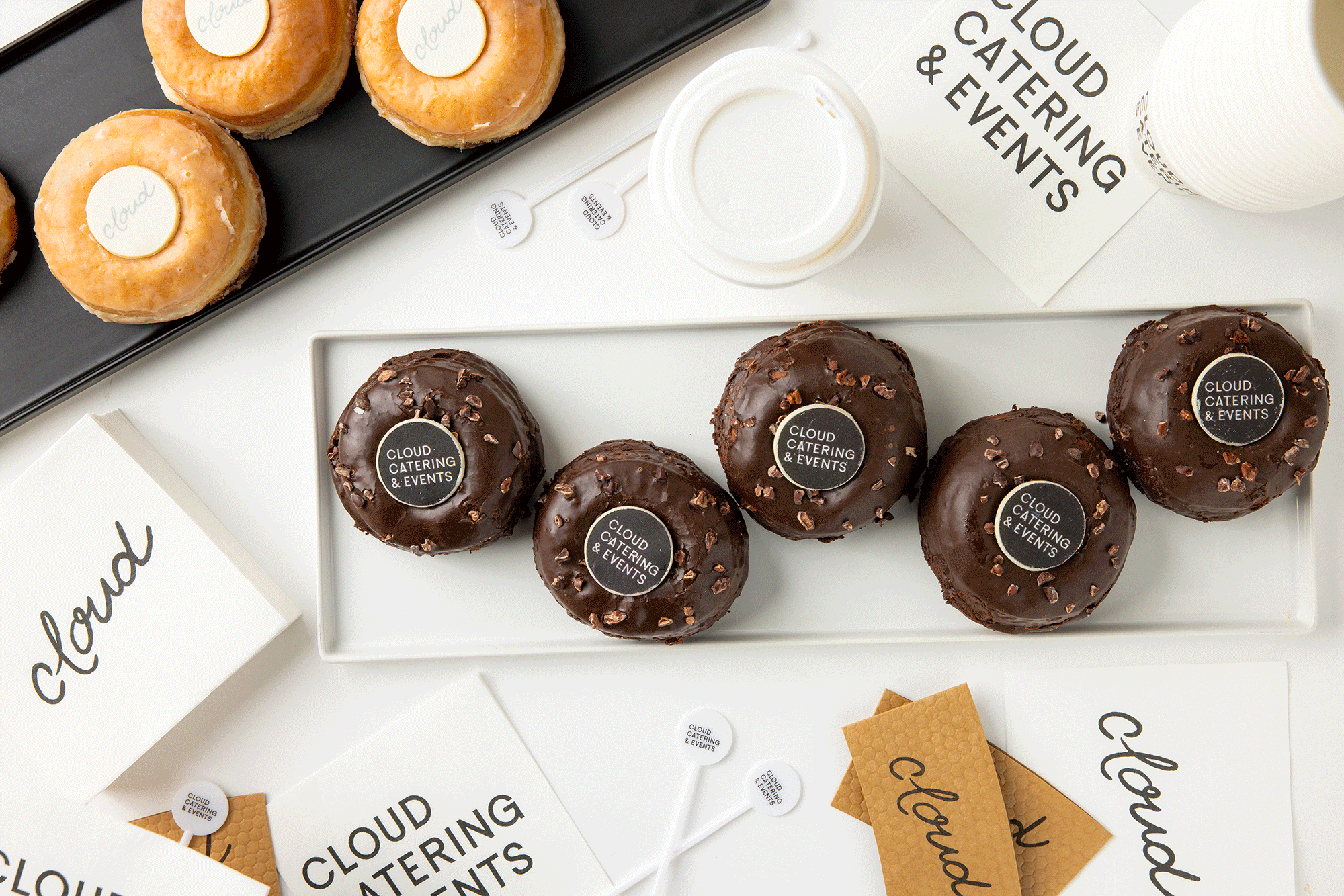 Assorted miniature desserts on a white and black surface, including chocolate-glazed donuts with 'Cloud Catering & Events' labels and round glazed pastries with white and brown layers, along with name cards, paper cups, and lollipops with the same lo