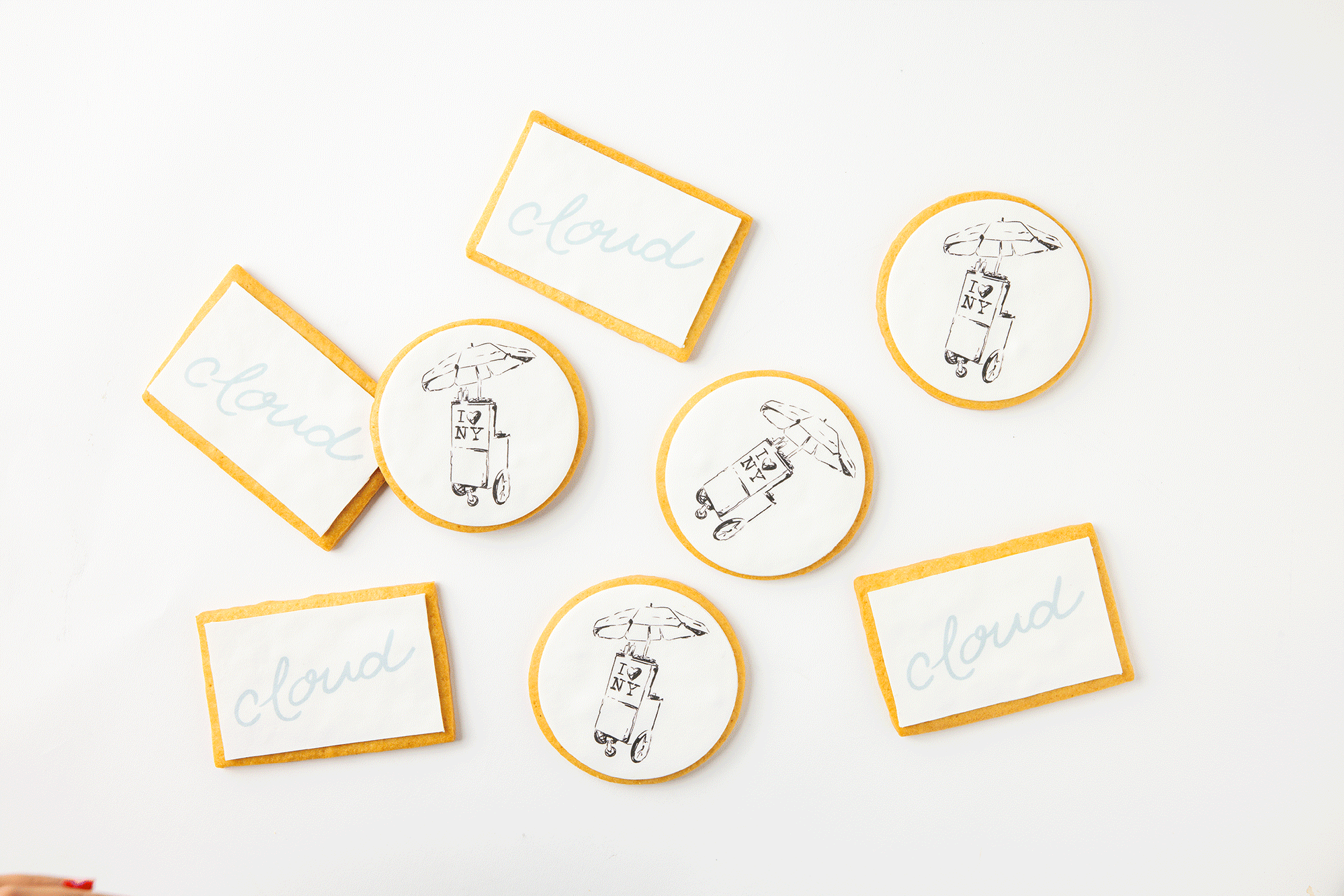 Decorative cookies with designs and text, some with the word 'cloud' and others with an illustration of a luggage cart with an umbrella and 'I ❤️ NY' sign, scattered on a white surface.