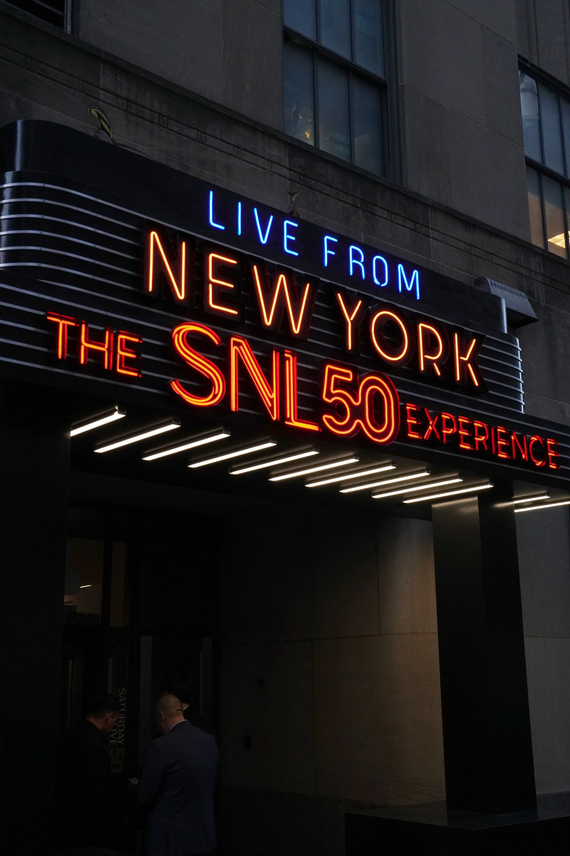 Neon sign reading 'Live from New York, the SNL 50 Experience' outside a building at night. Two people are standing near the entrance.