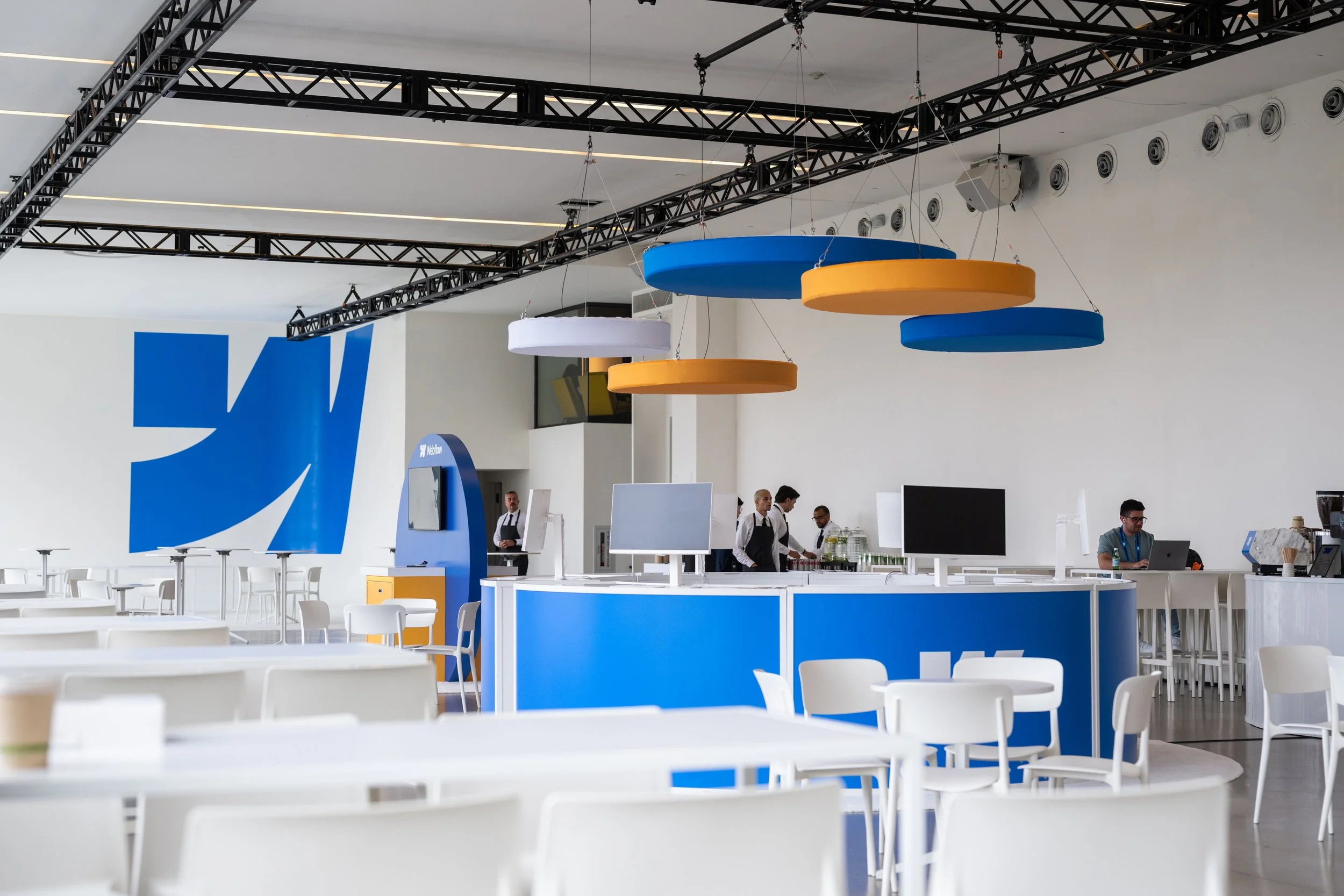 Modern cafe with white tables and chairs, blue and orange circular hanging decorations, a blue counter with computer screens, and staff in uniforms. Large blue logo on the wall.