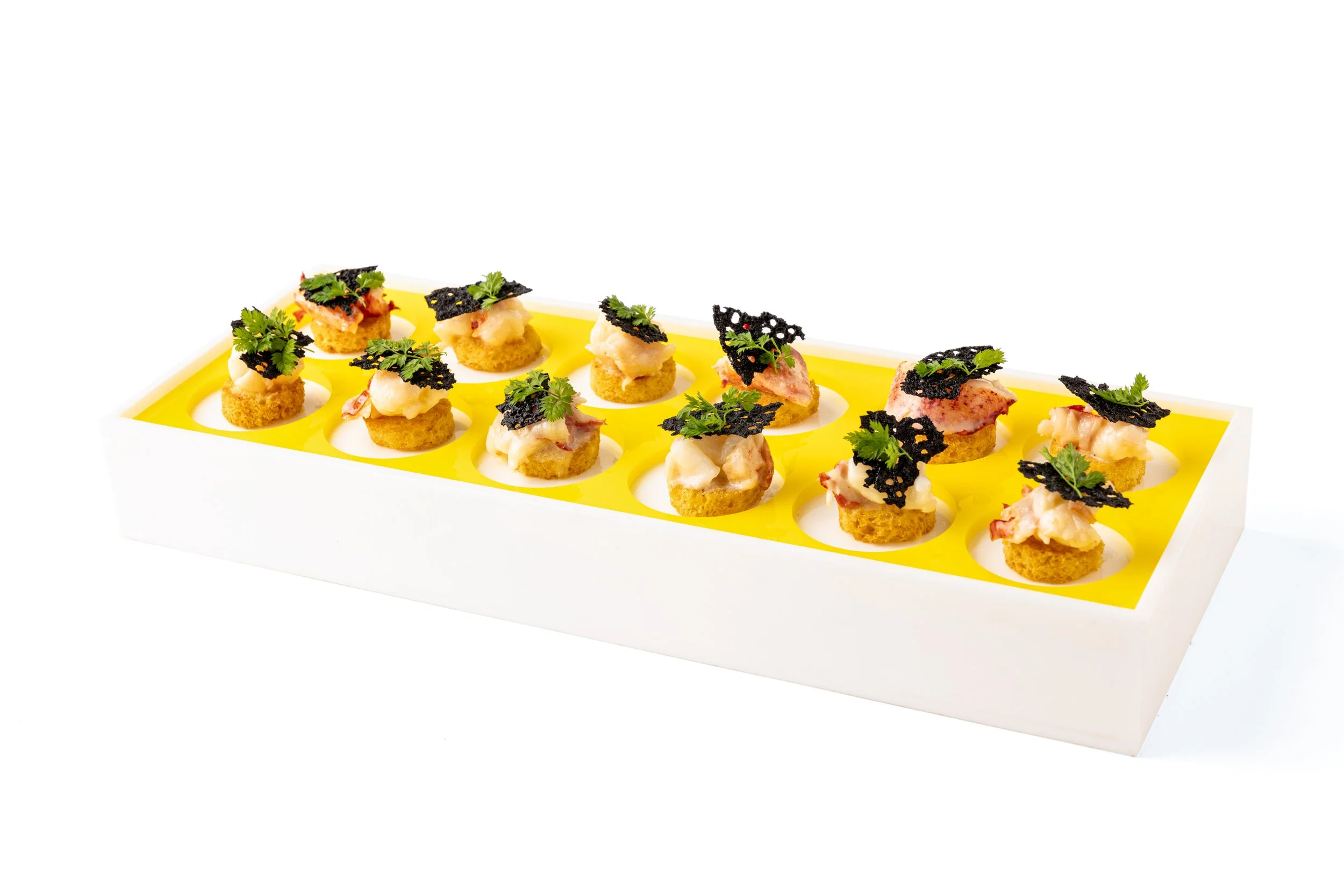 Elevated view of assorted canapé bites on a white and yellow serving tray, topped with herbs and black crisp garnish.