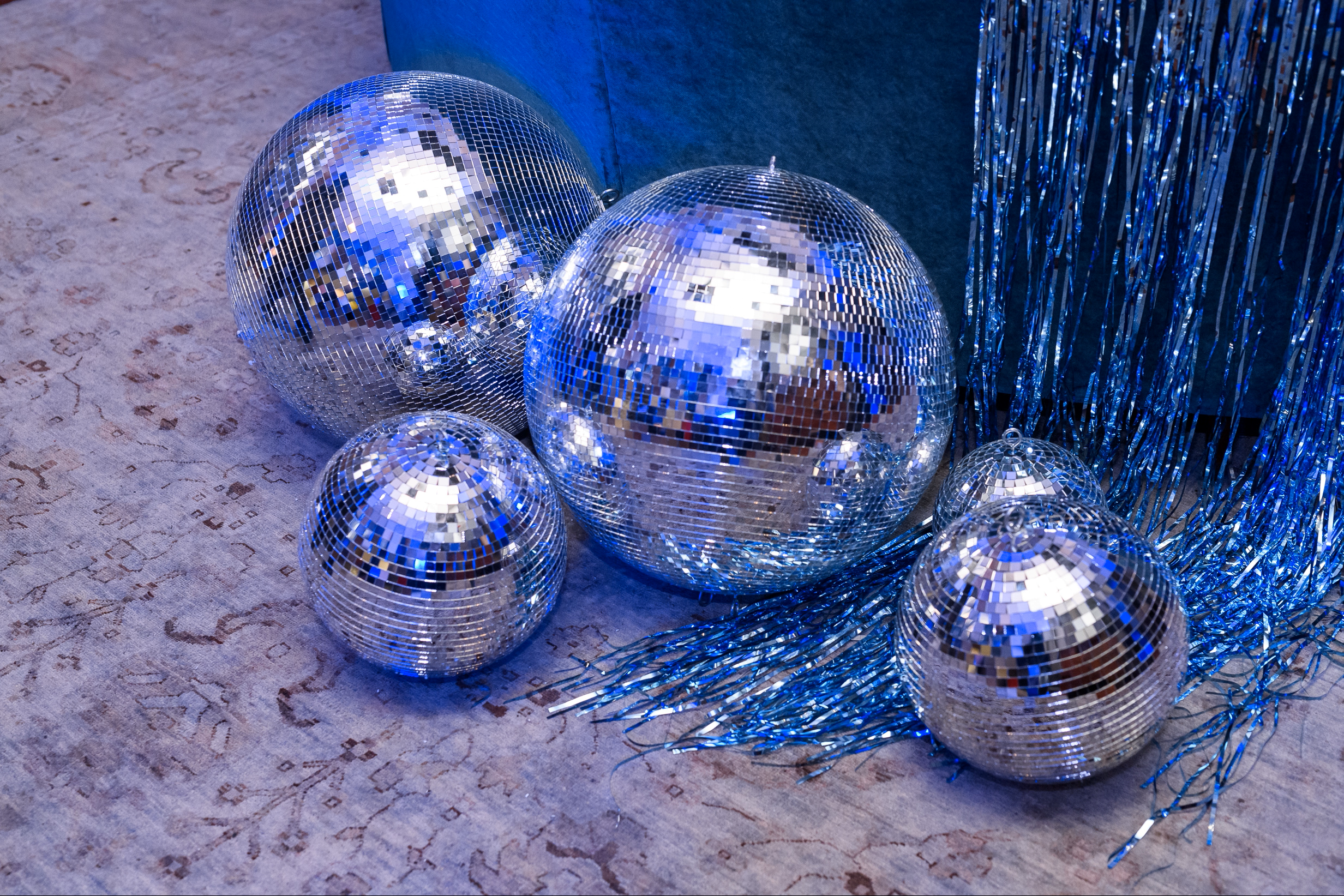 A collection of six disco balls of various sizes with reflective mirrored surfaces, placed on a textured floor with blue tinsel garland nearby, against a blue wall background.