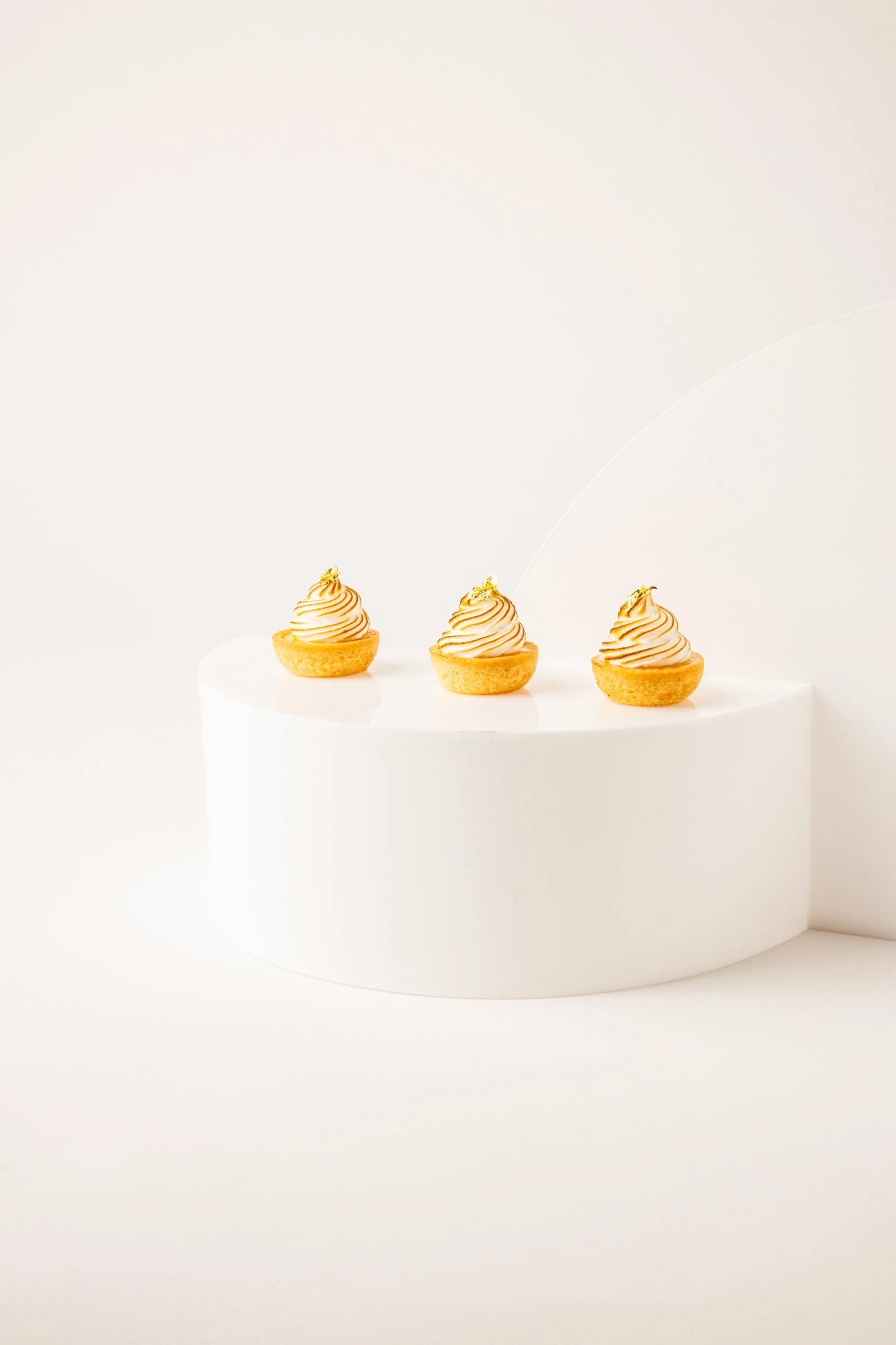 Three small, individual tart desserts with piped whipped cream and a gold leaf, displayed on a white, semi-circular platform against a plain white background.
