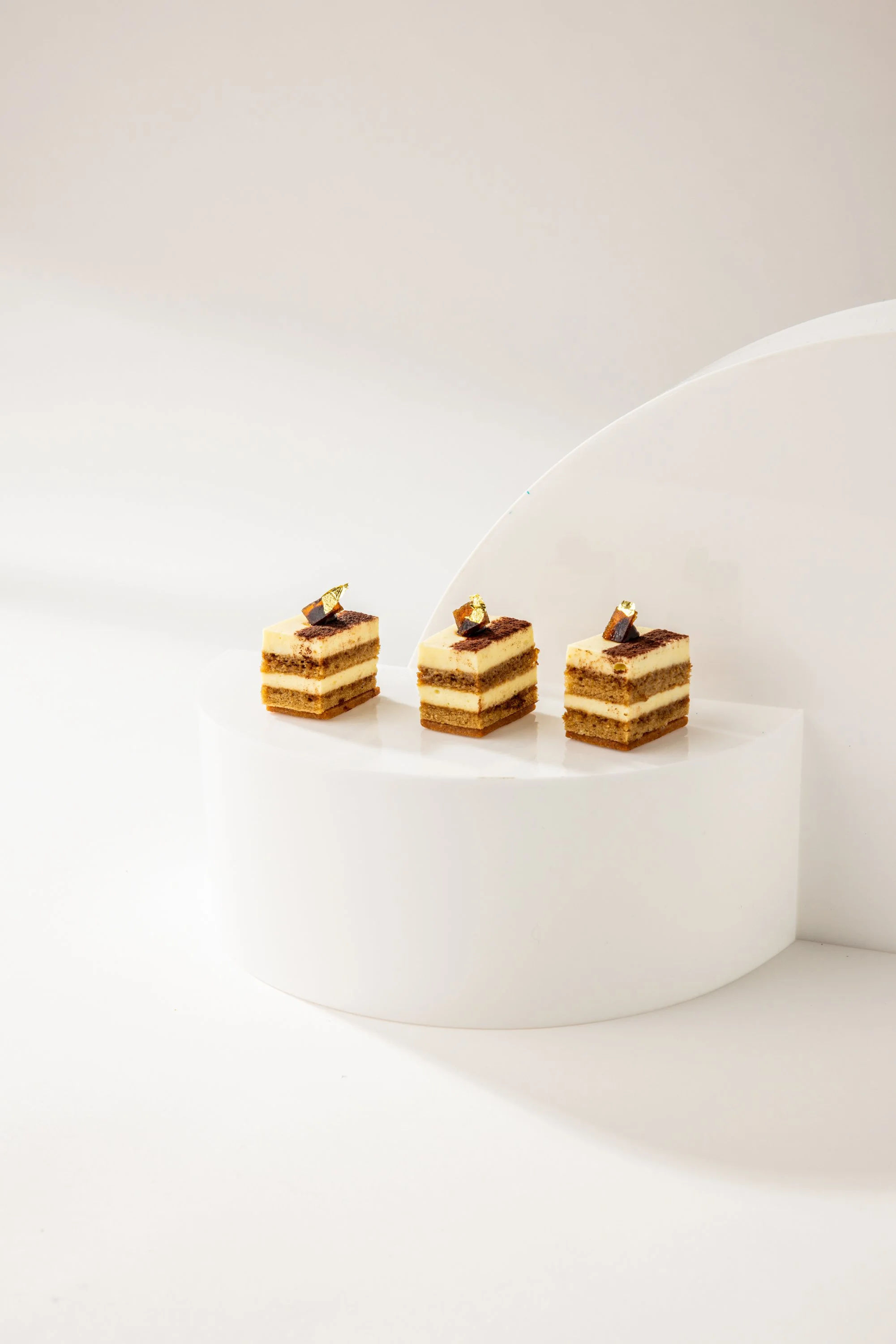 Three small layered desserts topped with gold leaf pieces on a white pedestal against a white background.