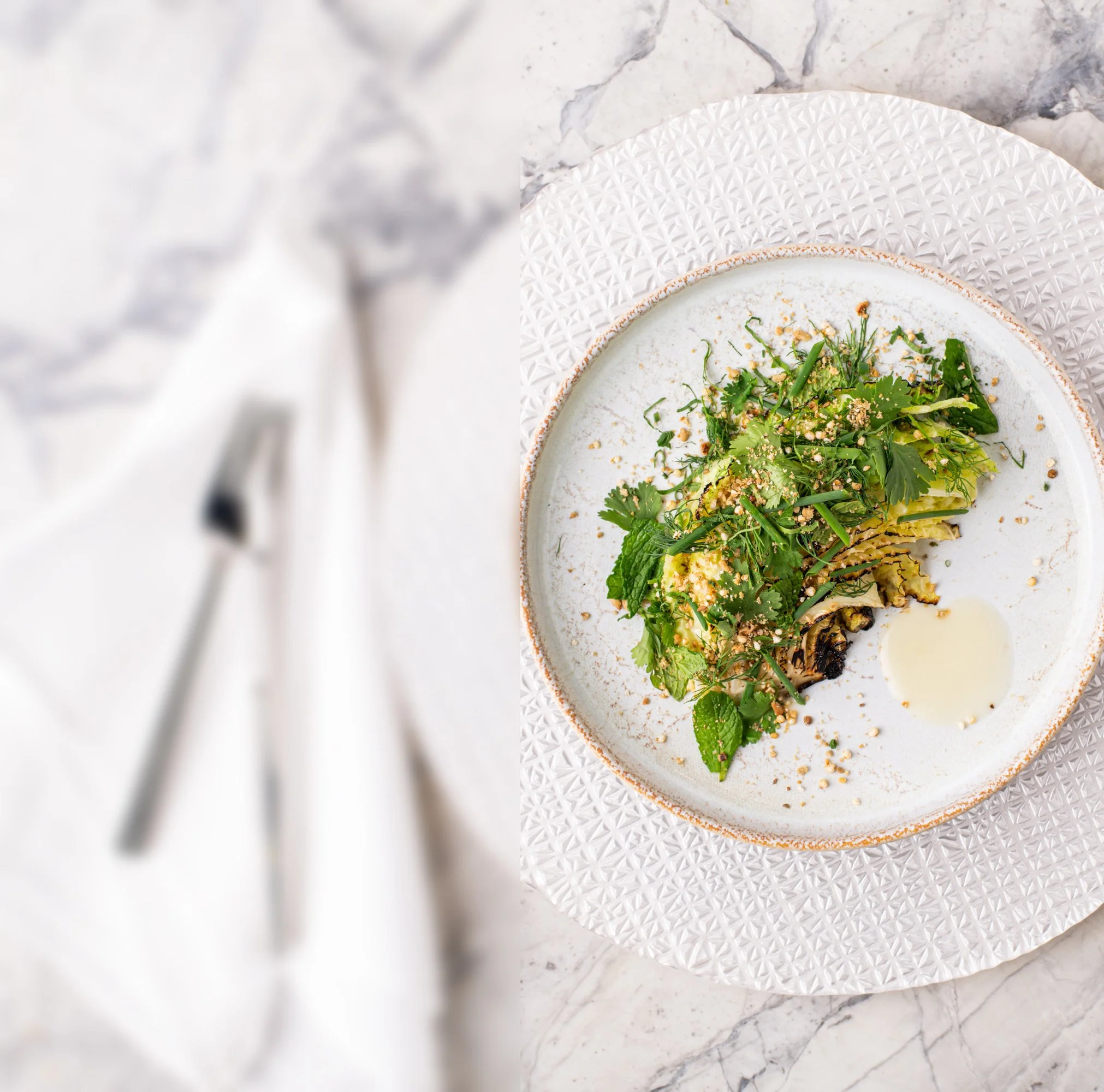 A white plate with a textured rim holds a fresh green salad with herbs, greens, sesame seeds, and a drizzle of dressing, on a white surface with a marbled background.