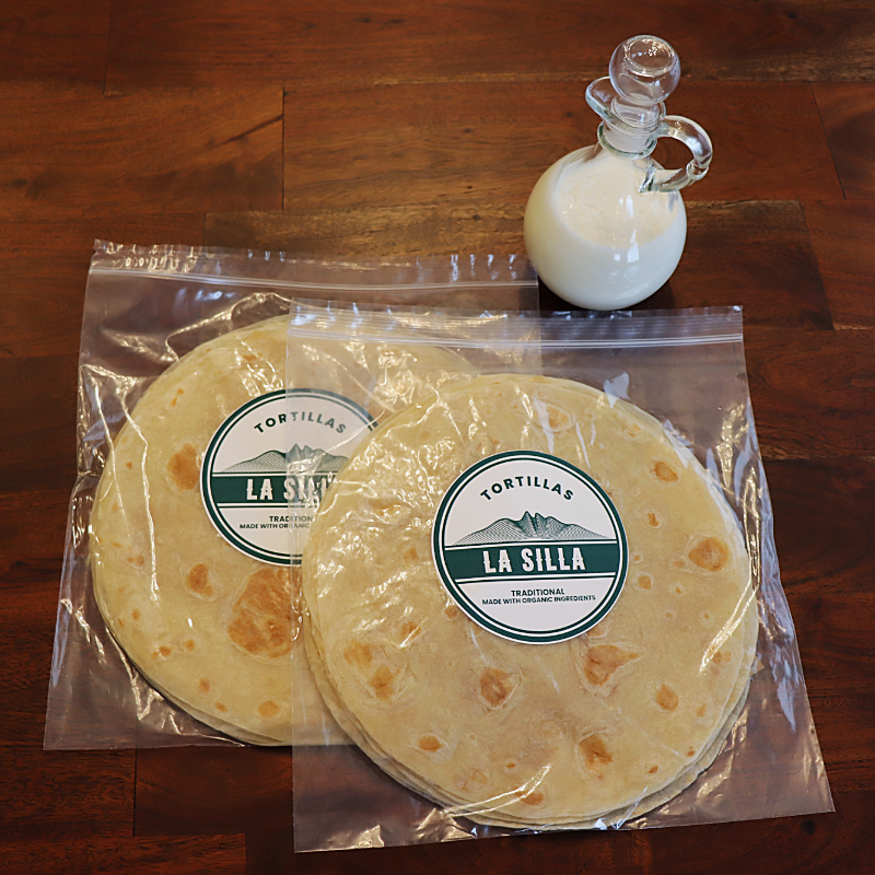 Two packaged tortillas on a wooden surface with a small glass pitcher of milk or cream nearby.