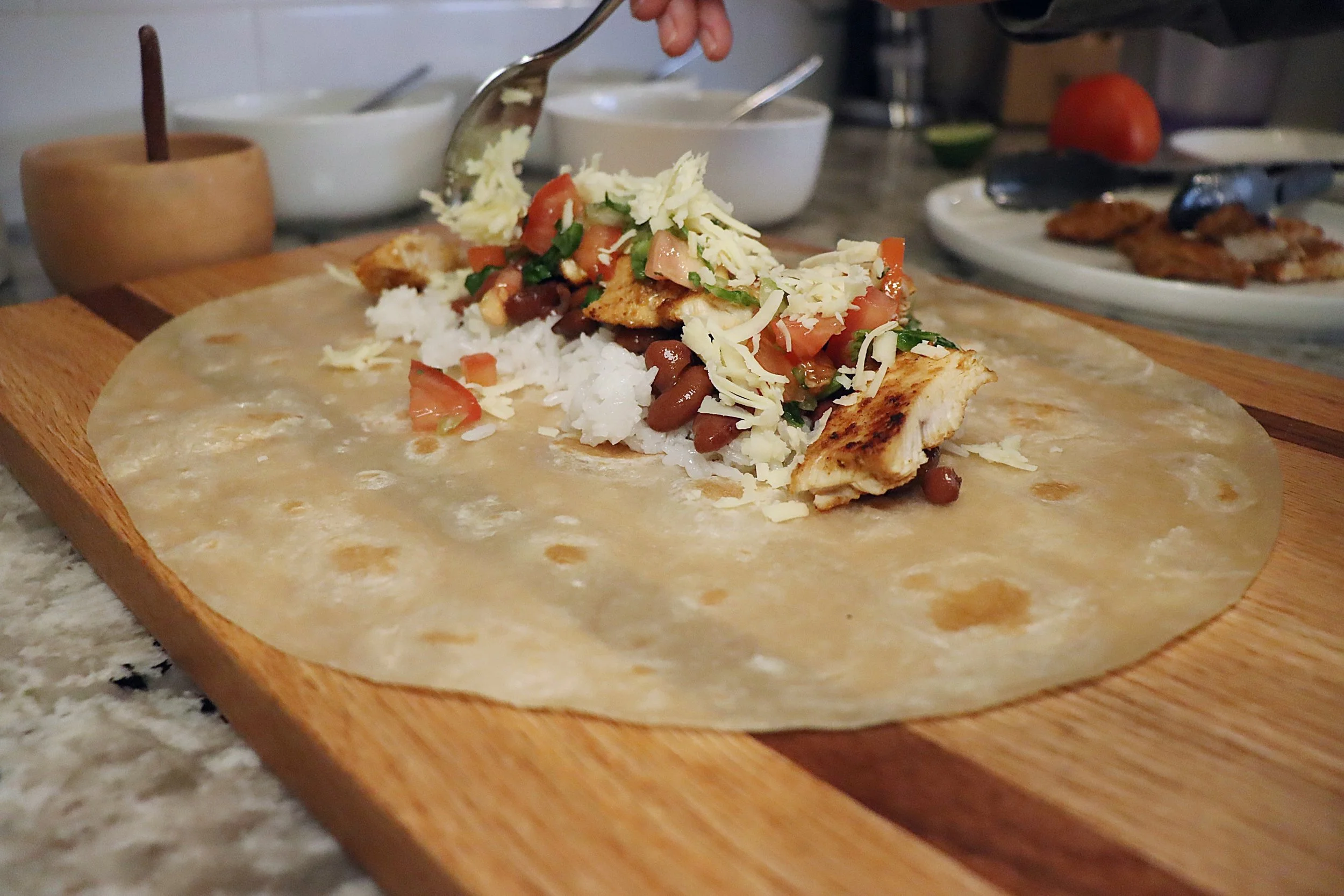 Preparation of a taco with grilled chicken, tomatoes, beans, cheese, shredded lettuce, and rice on a flour tortilla, placed on a wooden cutting board.