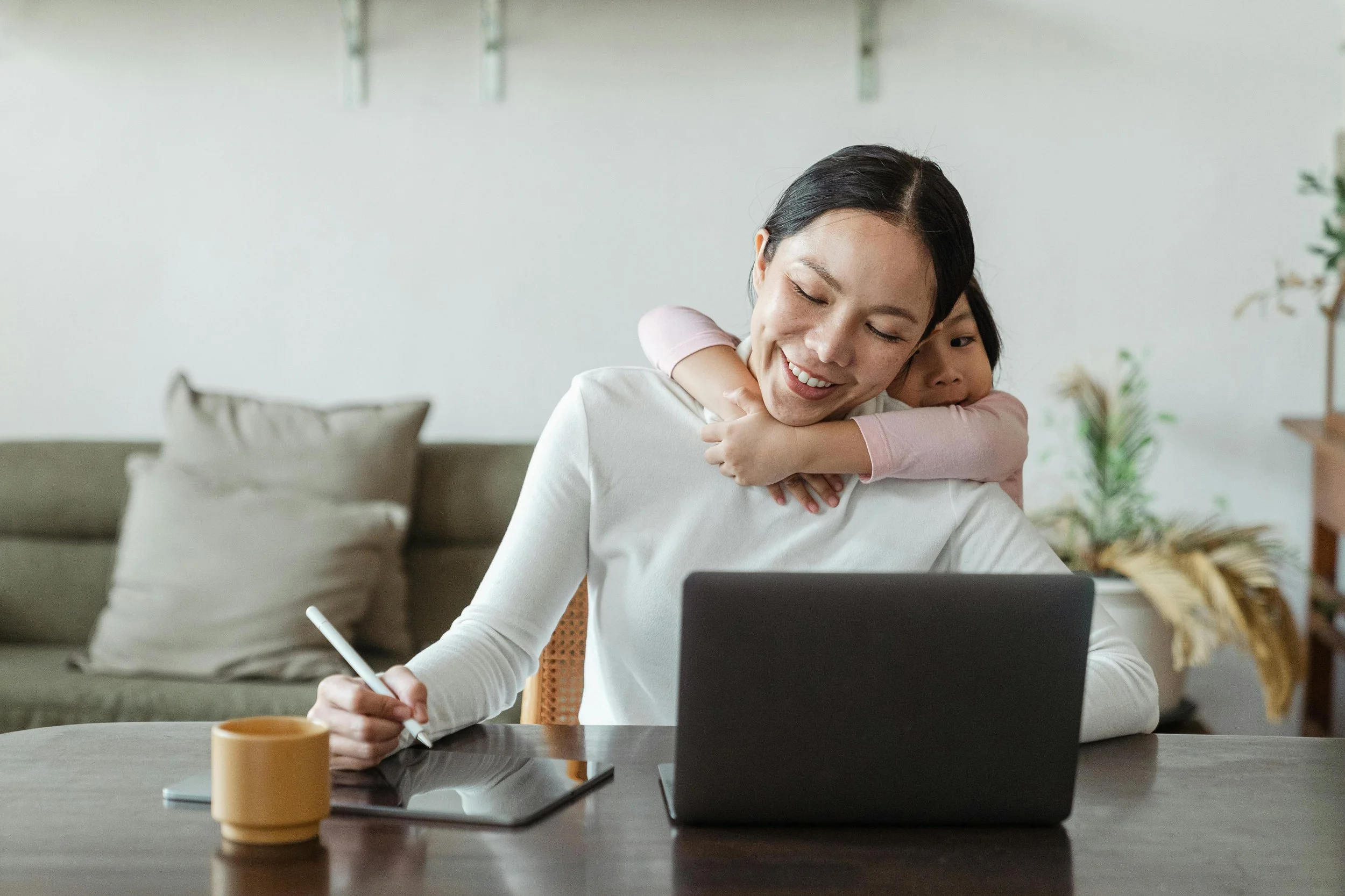 A woman sitting at a desk with a laptop, holding a stylus on a graphics tablet, and a young girl hugging her from behind in a cozy home setting.