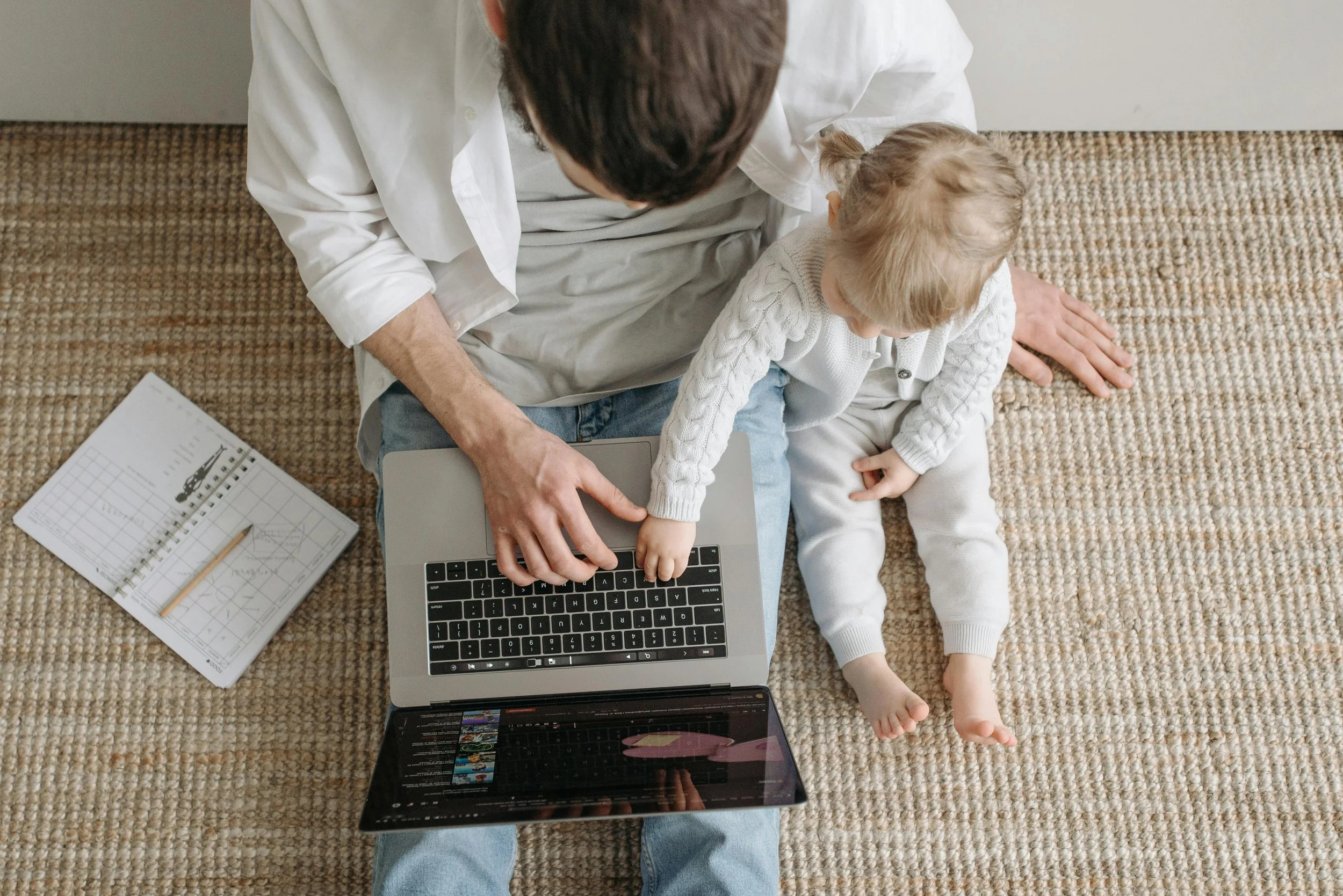 man typing on laptop next to child sitting on the floor with an open notepad next to him