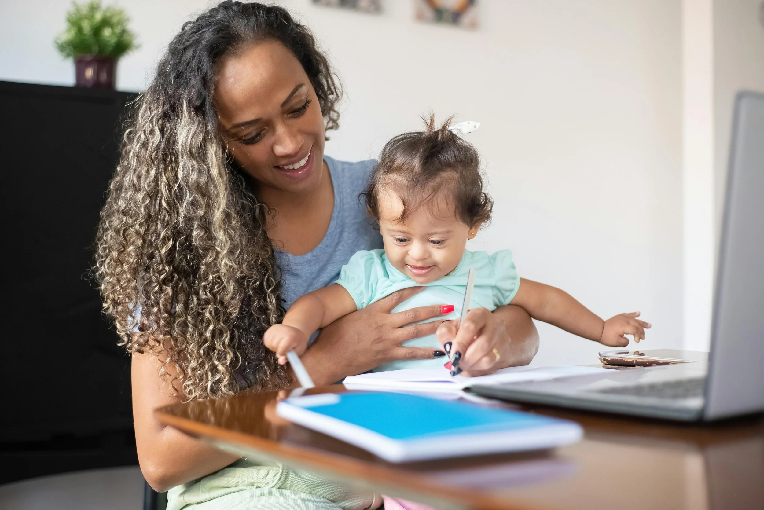 A woman with long, curly hair holds a young girl on her lap as they write together at a desk with a laptop, notebooks, and papers in a home setting.