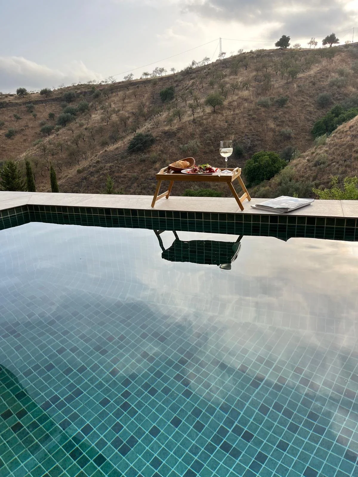 Schwimmbad mit Blick auf hügelige Landschaft mit Bäumen, Picknicktablett mit Wein, Salat, Brot und Gläsern