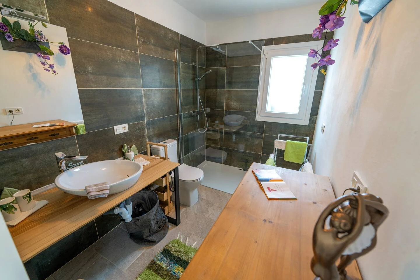 Ein Badezimmer mit dunklen Wandfliesen, einer Dusche mit Glasabtrennung, einem Fenster, einem weißen Waschbecken auf einem Holzregal, einem Spiegel, einem Toilettensitz, Handtüchern und Pflanzen.