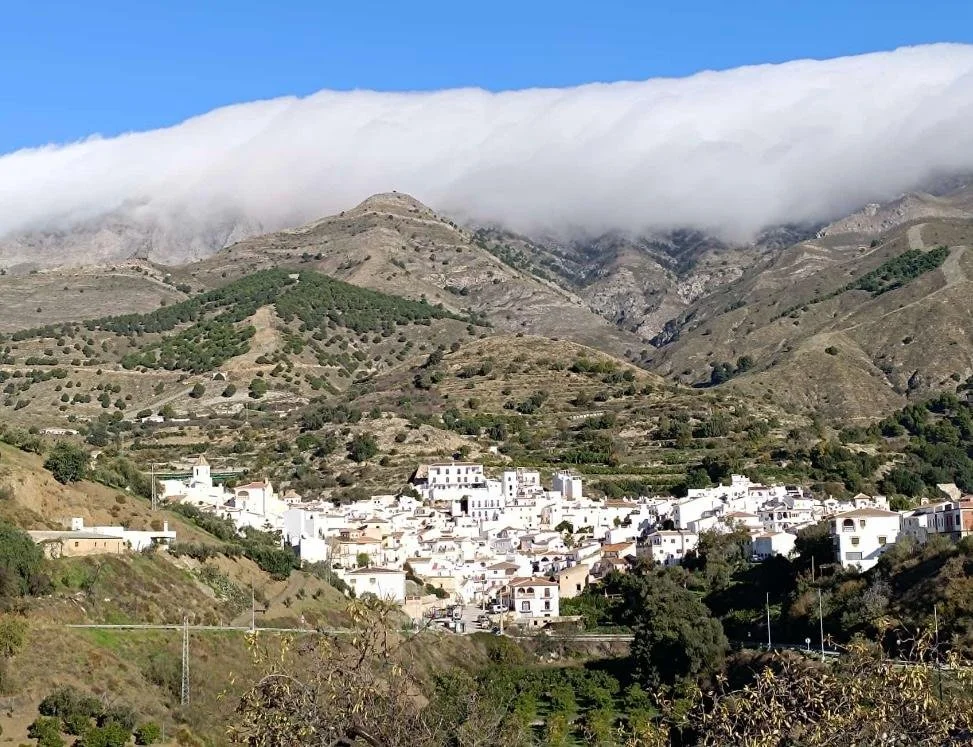 Kleines Dorf mit weißen Gebäuden inmitten von grünen Hügeln und Bergen, Wolken hängen über den Bergen.