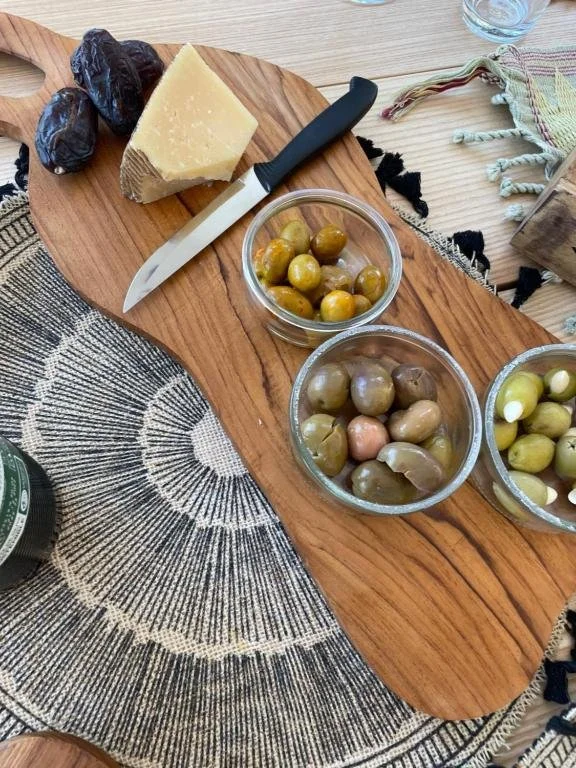 Verschiedene Oliven in kleinen Gläsern, ein Stück Hartkäse, Feigen, ein Messer mit schwarzem Griff, auf einem Holzbrett, daneben ein Glas Wasser und eine bunte Stoffserviette.