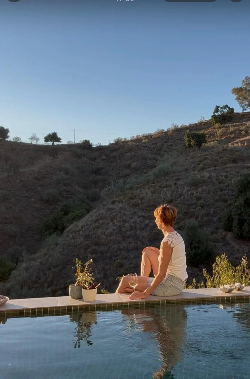 Eine Frau sitzt am Rand eines Swimmingpools, blickt auf eine bergige Landschaft bei Sonnenuntergang, mit einem Glas Wein in der Hand.