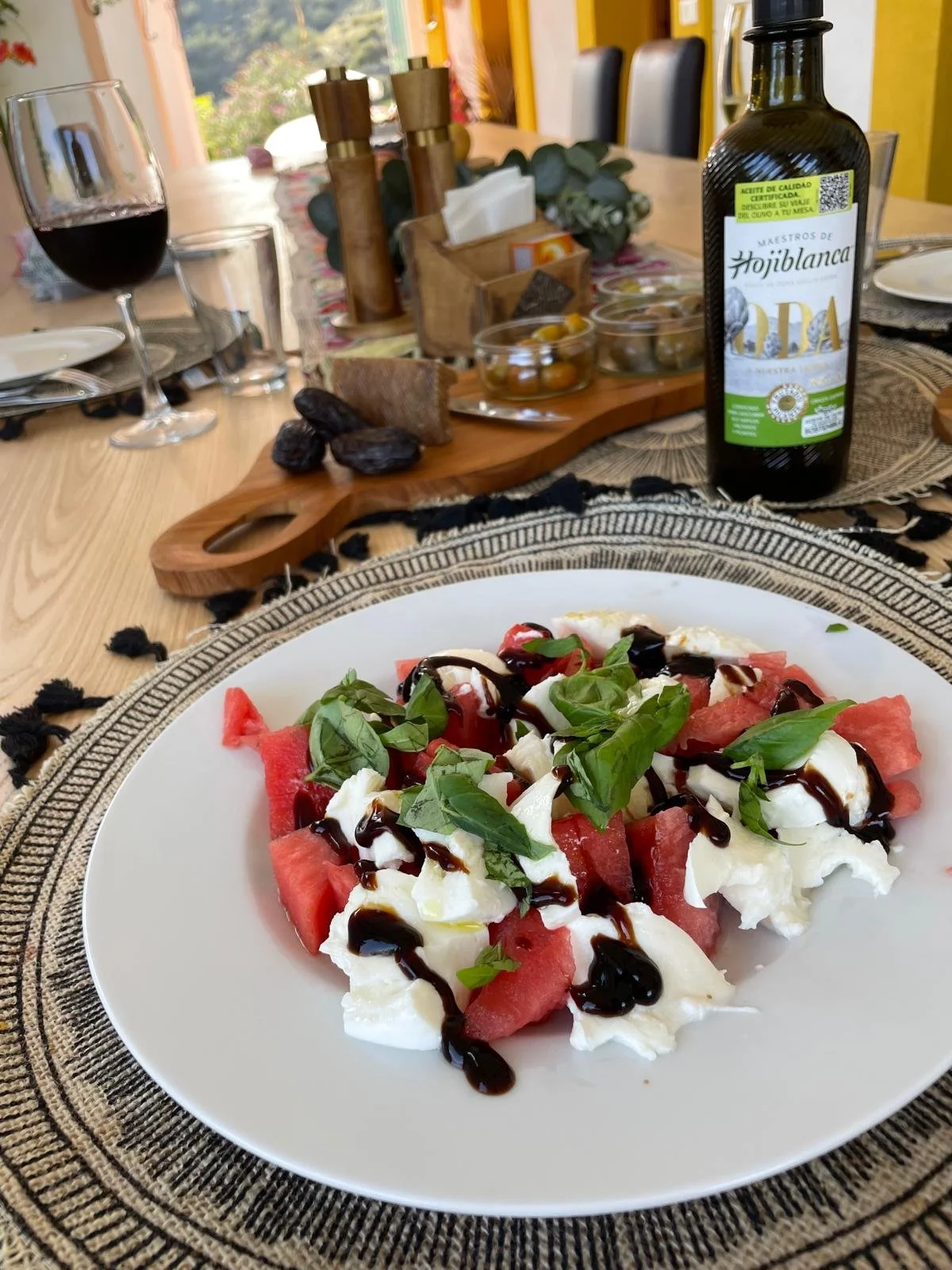 Salat mit Wassermelone, Mozzarella, frischen Basilikumblättern und Balsamico-Reduktion auf einem weißen Teller, im Hintergrund Wein, Brot, Olivenöl, und Gewürze auf einem Holzteller.
