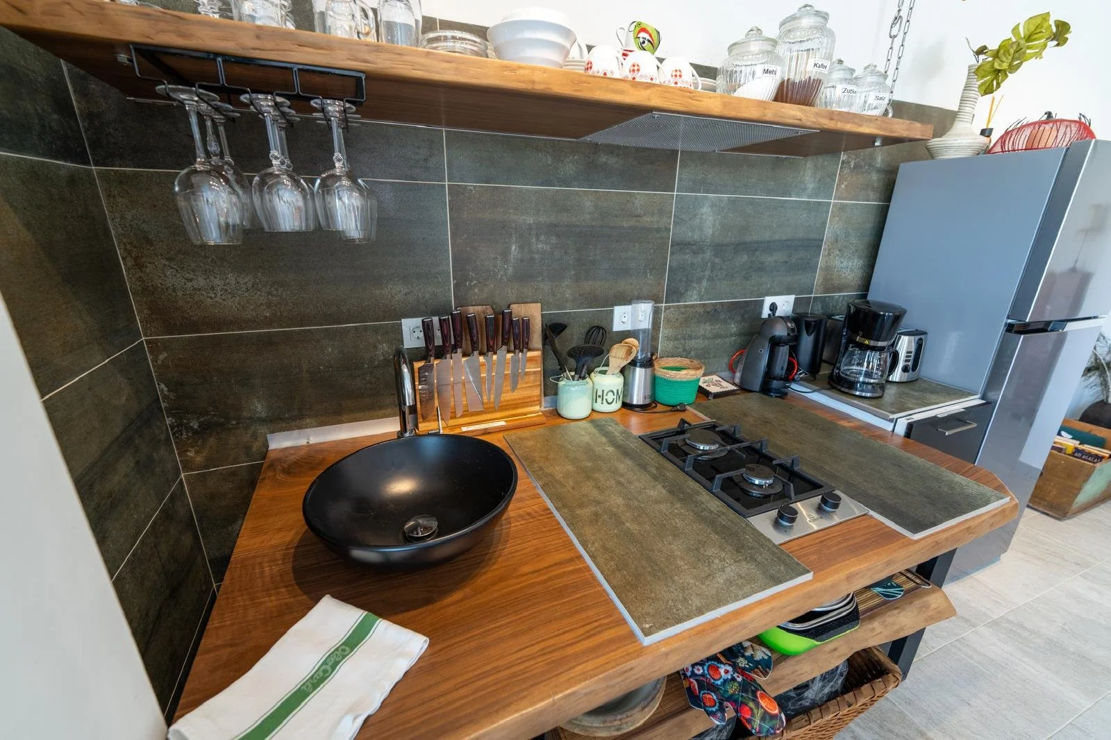 Küchenarbeitsplatte mit schwarzen Waschbecken, Messerblock, Kaffeekanne, Kaffeemaschine, Kühlschrank, Gläser, Vorratsgläser, Küchenutensilien und Geschirr, offene Regale