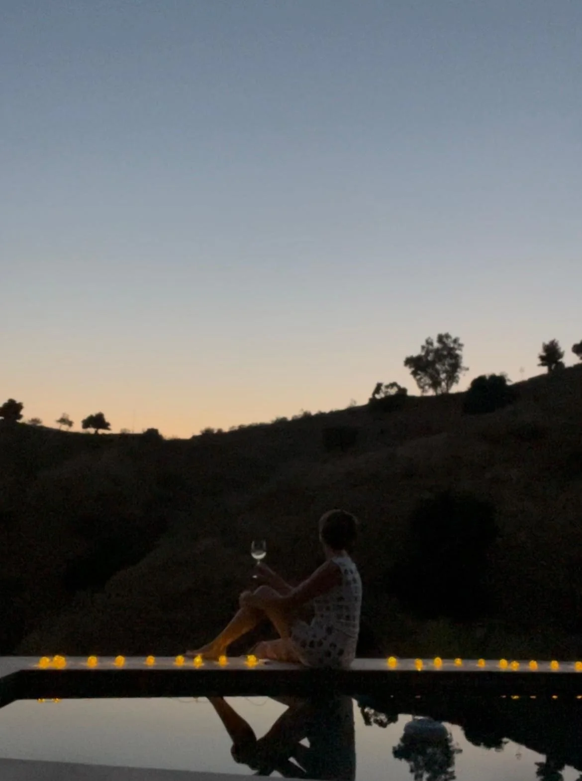Simone Wittig sitzt an einem sonnigen Abend auf einer Brücke mit Lichterketten, hält ein Glas und schaut auf die Berge und Bäume im Hintergrund.