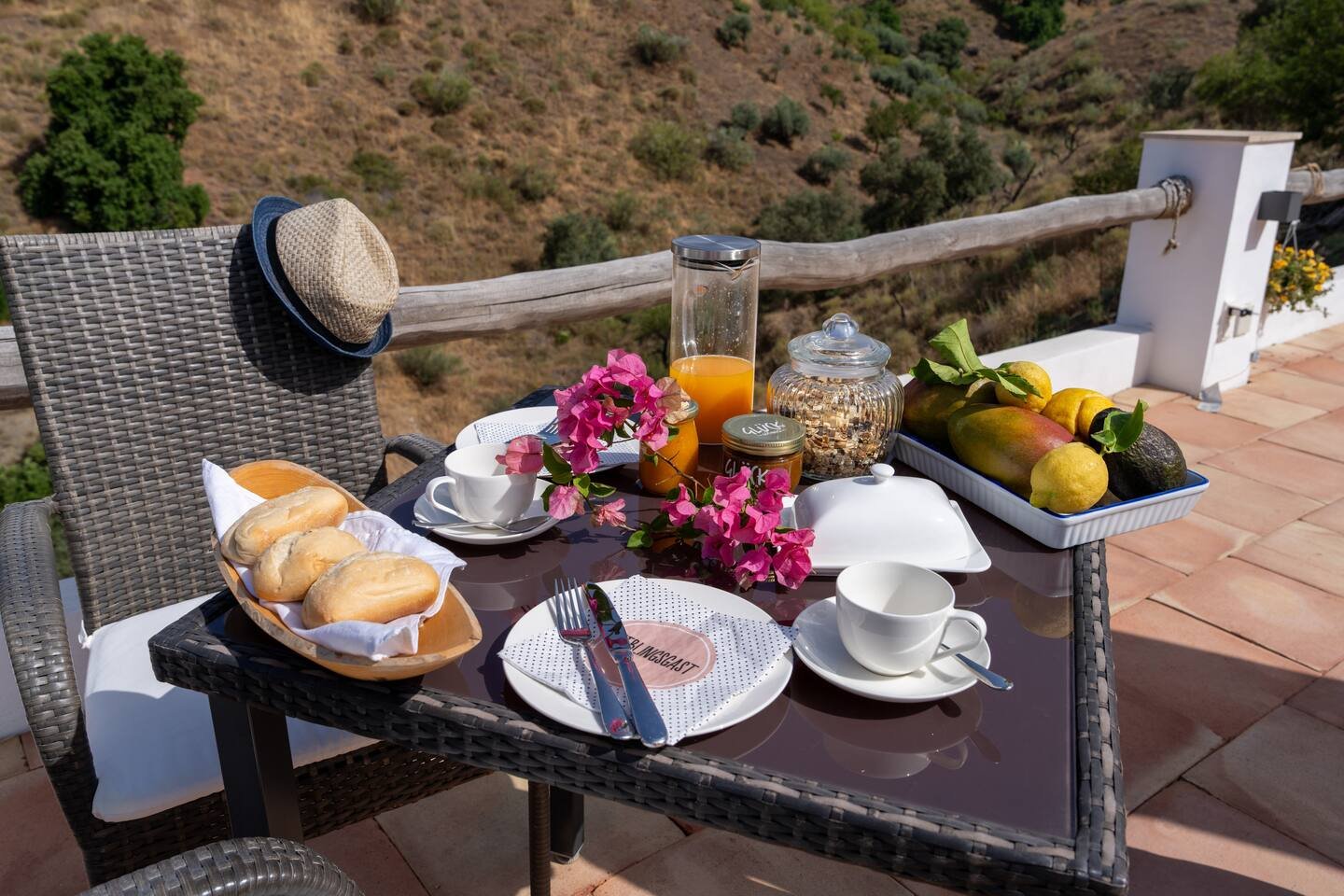 Ein Frühstückstisch im Freien auf einer Terrasse mit Blick auf eine hügelige Landschaft, gedeckt mit Broten, Obst, Kaffee, Tee und Orangensaft, dekoriert mit Blumen.