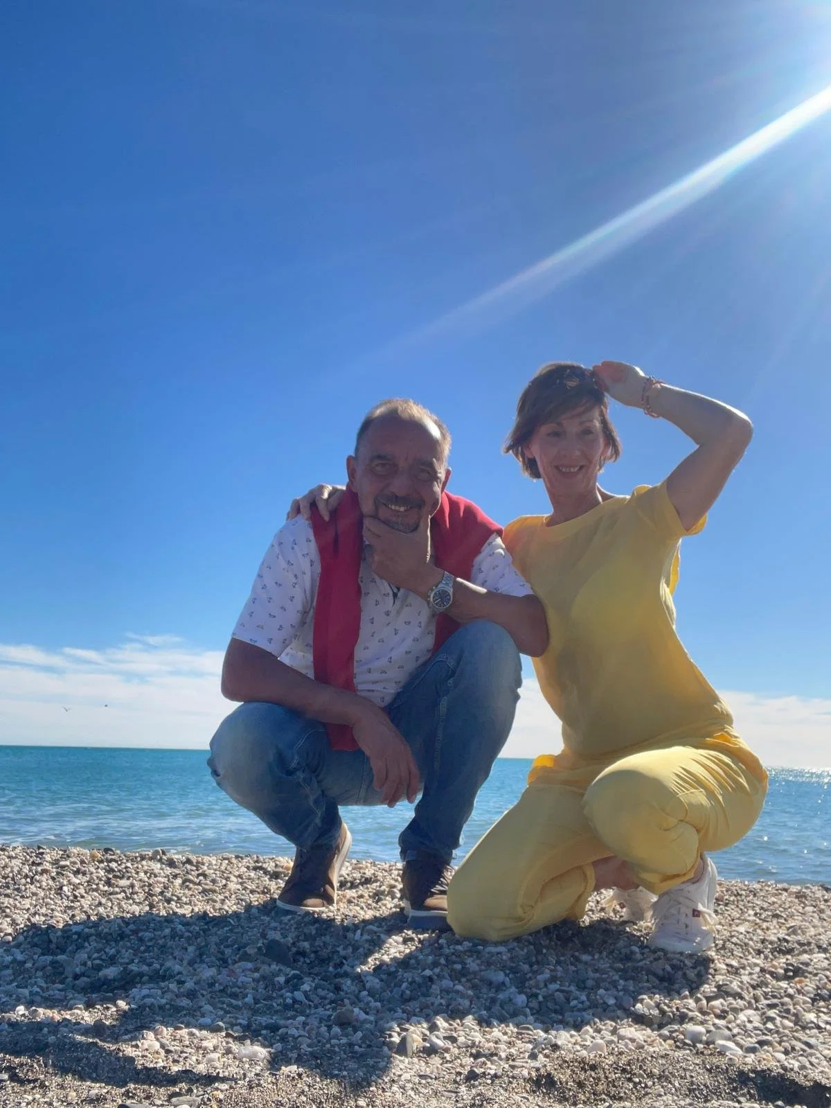 Zwei Menschen, ein Mann und eine Frau, posieren am Strand bei Sonnenschein mit dem Meer im Hintergrund.