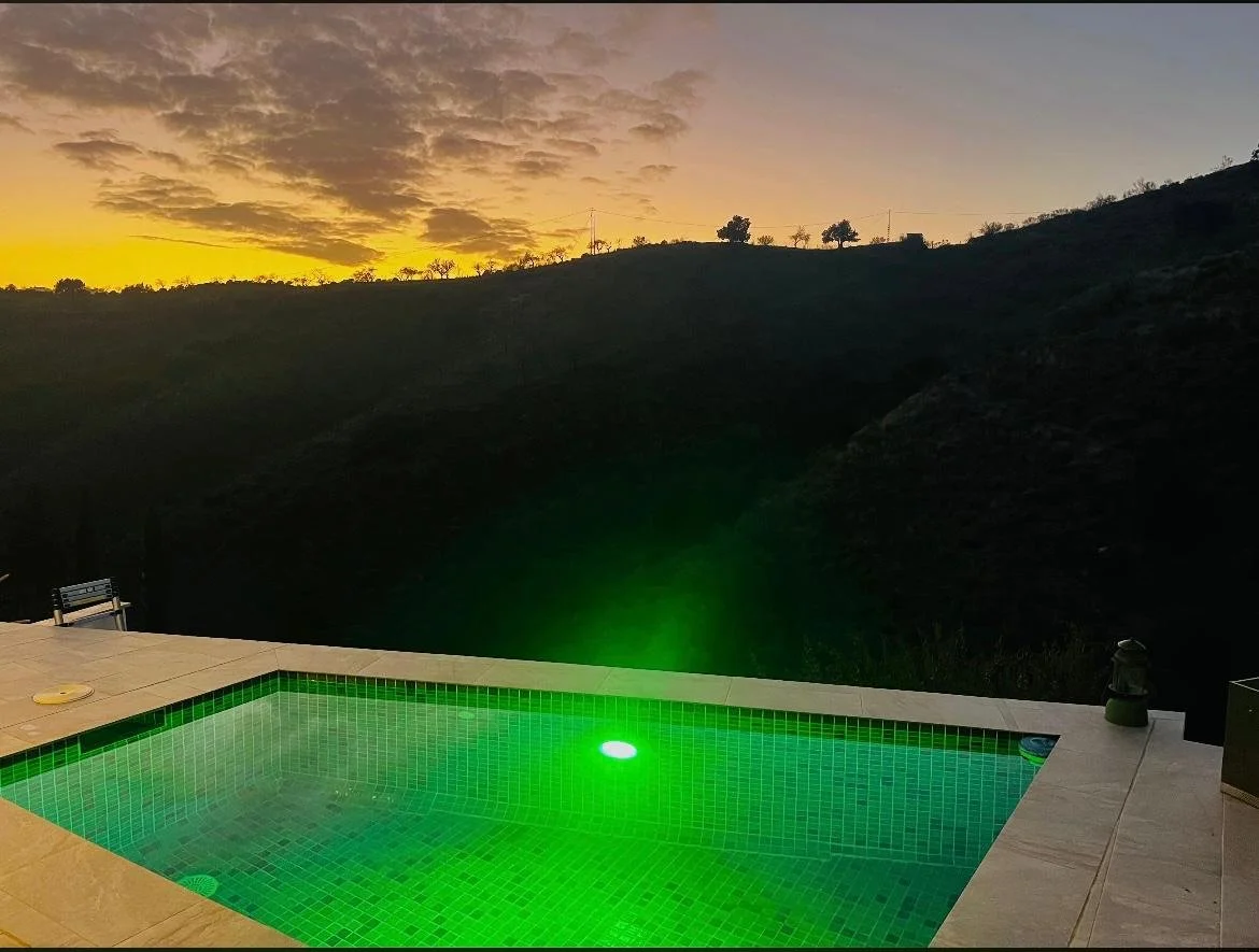 Ein Pool mit grüner Beleuchtung im Vordergrund. Im Hintergrund ein Berg und ein Himmel bei Sonnenuntergang