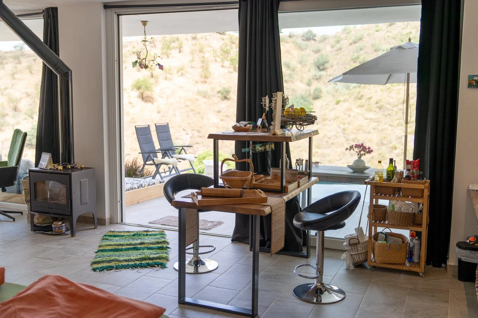 Innenansicht eines Wohnzimmers mit Esstisch, Barhockern, einem Regal mit Küchenutensilien, einem Kamin, Vorhängen und Blick auf einen Balkon mit Liegestühlen und einem Sonnenschirm im Freien