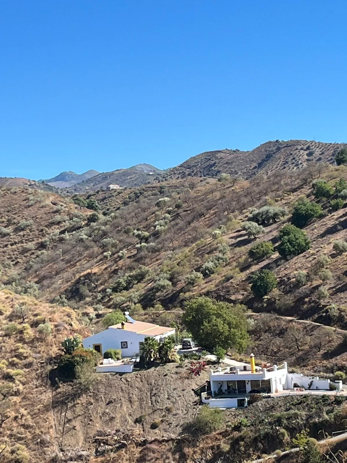 Blick auf ländliche weiße Häuser auf hügeligem, trockenen Gelände mit wenigen Bäumen, vor Bergkette und klarem blauen Himmel.