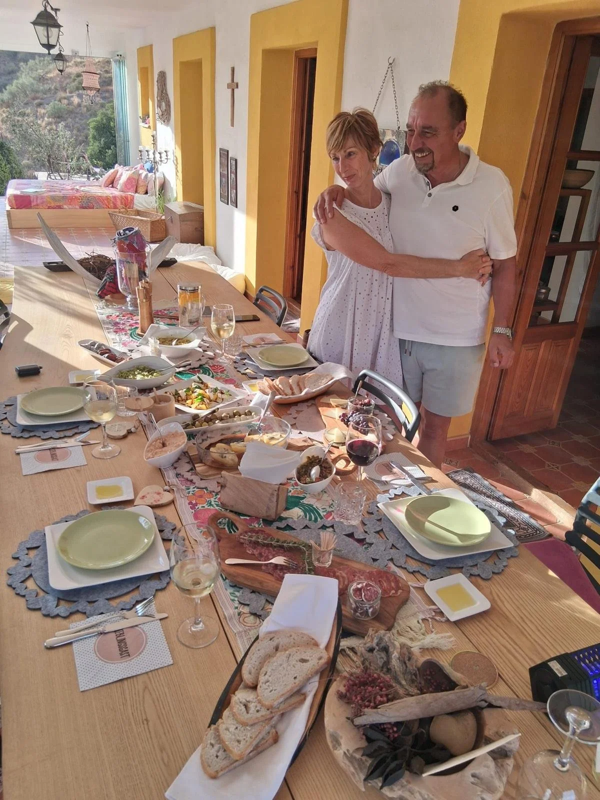 Ein festlich gedeckter Esstisch mit verschiedenen Speisen, Wein und Wein glasses, in einem hellen Raum mit zwei Menschen, die sich umarmen.