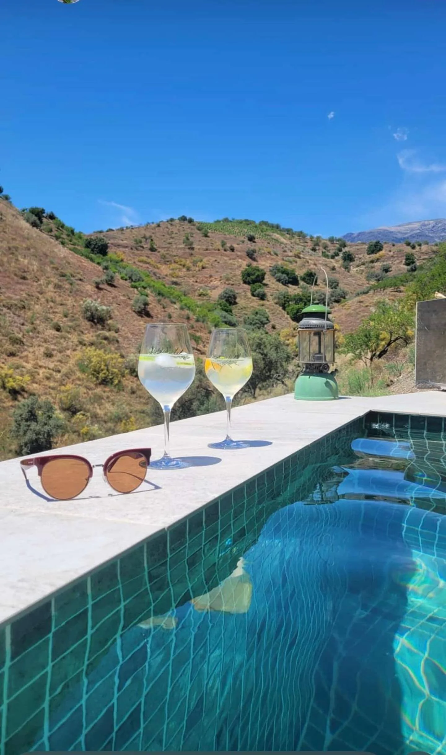 Sonnige Szenerie mit einem Swimmingpool im Vordergrund, zwei Gläser mit Wasser und Zitronenscheiben, eine Sonnenbrille auf der Poolkante, im Hintergrund hügelige Landschaft und blauer Himmel.