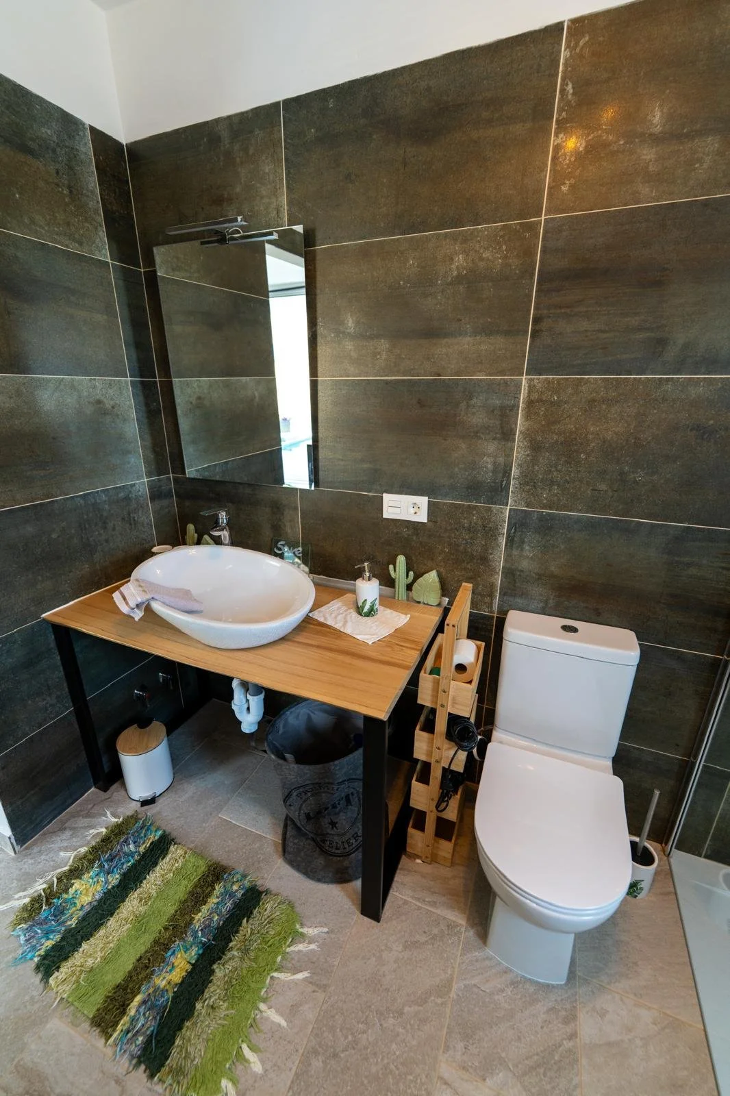 Modernes Badezimmer mit schwarzen Fliesen, weißem WC, Holz-Waschtisch mit rundem Waschbecken, Spiegel, Dekorationen wie Cactus und Seifenspender, bunte Teppichmatte, Toilettenpapierhalter und Hygieneartikel.