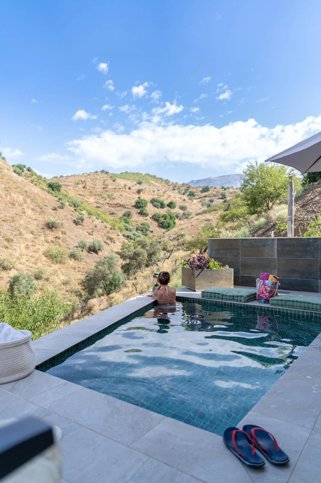 Eine Person entspannt im Infinity-Pool mit Blick auf bergige, trocken aussehende Landschaft und blauen Himmel