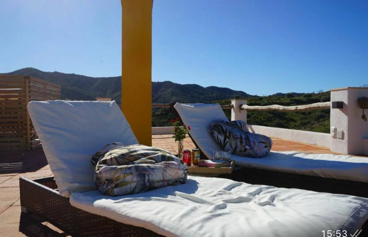 Sonnige Terrasse mit zwei Liegestühlen, einer mit einem gestreiften Kissen, Blick auf Hügel im Hintergrund, kleine Tische mit Getränken, Blumen und Sonnenschutz im Hintergrund.