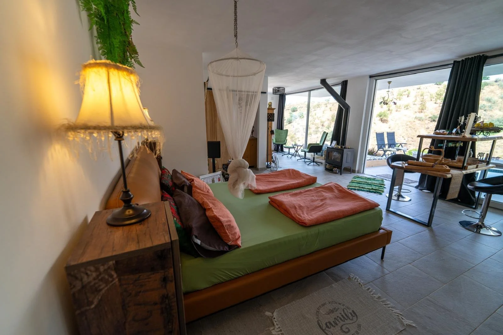 Gemütliches Wohnzimmer mit Doppelbett, Nachttischlampe, Blick auf Terrasse mit Stühlen und Garten, große Fenster, Sessel und Decor