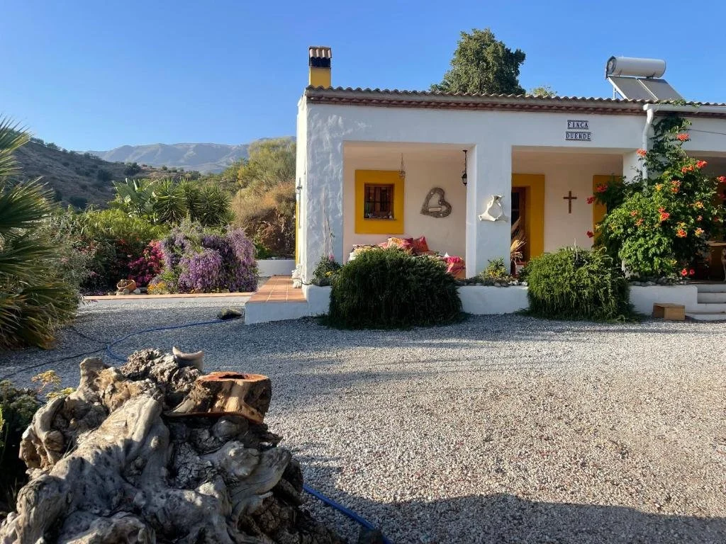 Ein weiames Haus mit einem kleinen Vorgarten, umgeben von Pflanzen und Sträuchern, mit Blumen und Dekorationen an der Wand, im Hintergrund Berge und blauer Himmel.