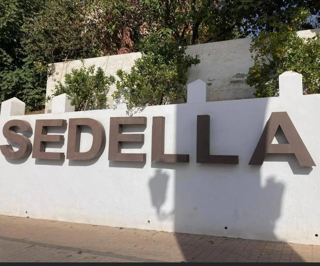 Wegweiser mit der Aufschrift 'SEDELLA' vor einer weißen Wand, mit Büschen und Bäumen im Hintergrund.