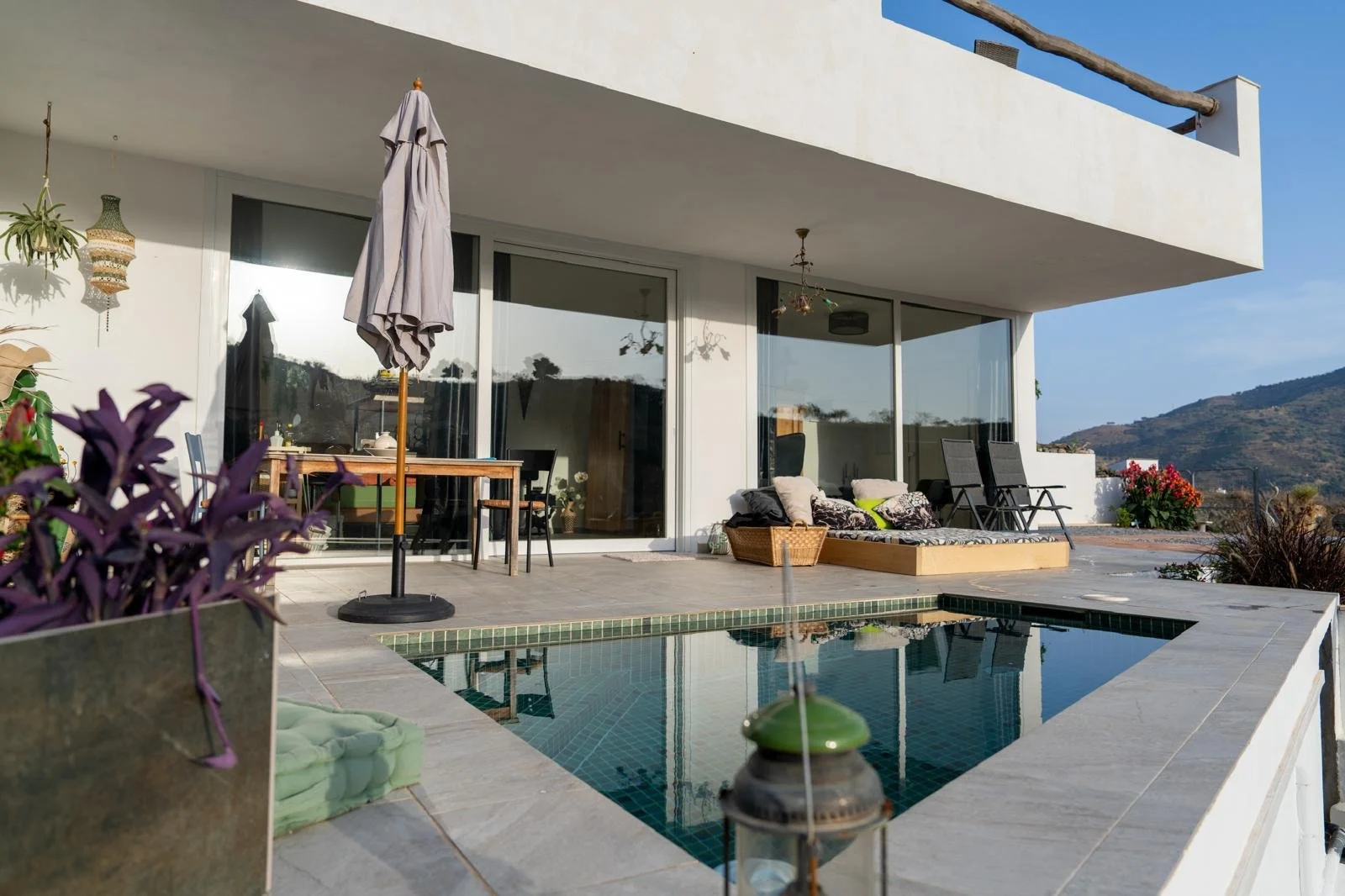 Geräumige Terrasse mit Pool, Sessel, Esstisch, Stühle, Sonnenschirm und Pflanzen an einem sonnigen Tag, mit Blick auf Berge im Hintergrund.