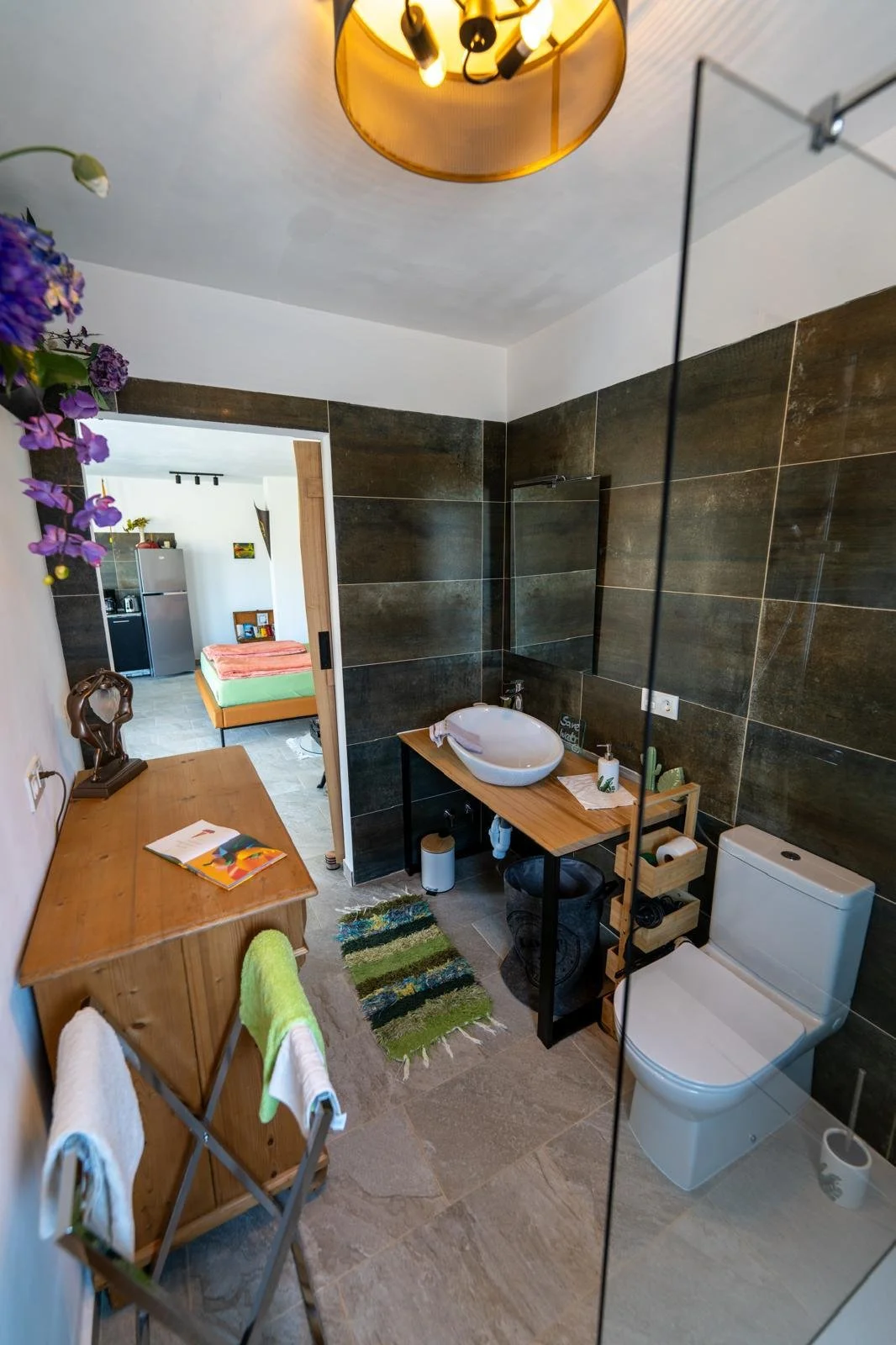 Badezimmer mit dunklen Wandfliesen, Waschbecken auf Holzregal, Toilette, Kleiderablage, kleine grüne Matte, Etagere mit Toilettenpapier, Handseife, Handtücher, dekorative Pendelleuchte, Blick in angrenzendes Schlafzimmer mit Bett und Kühlschrank.