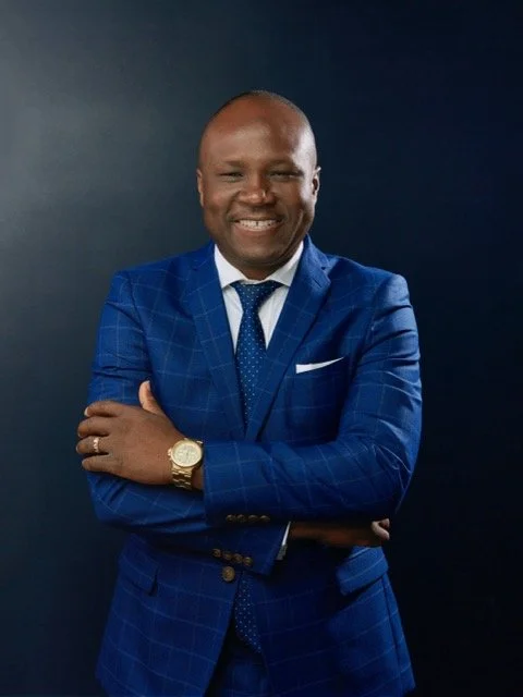 A smiling man in a blue checkered suit with a dress shirt and tie, standing against a dark background with arms crossed.