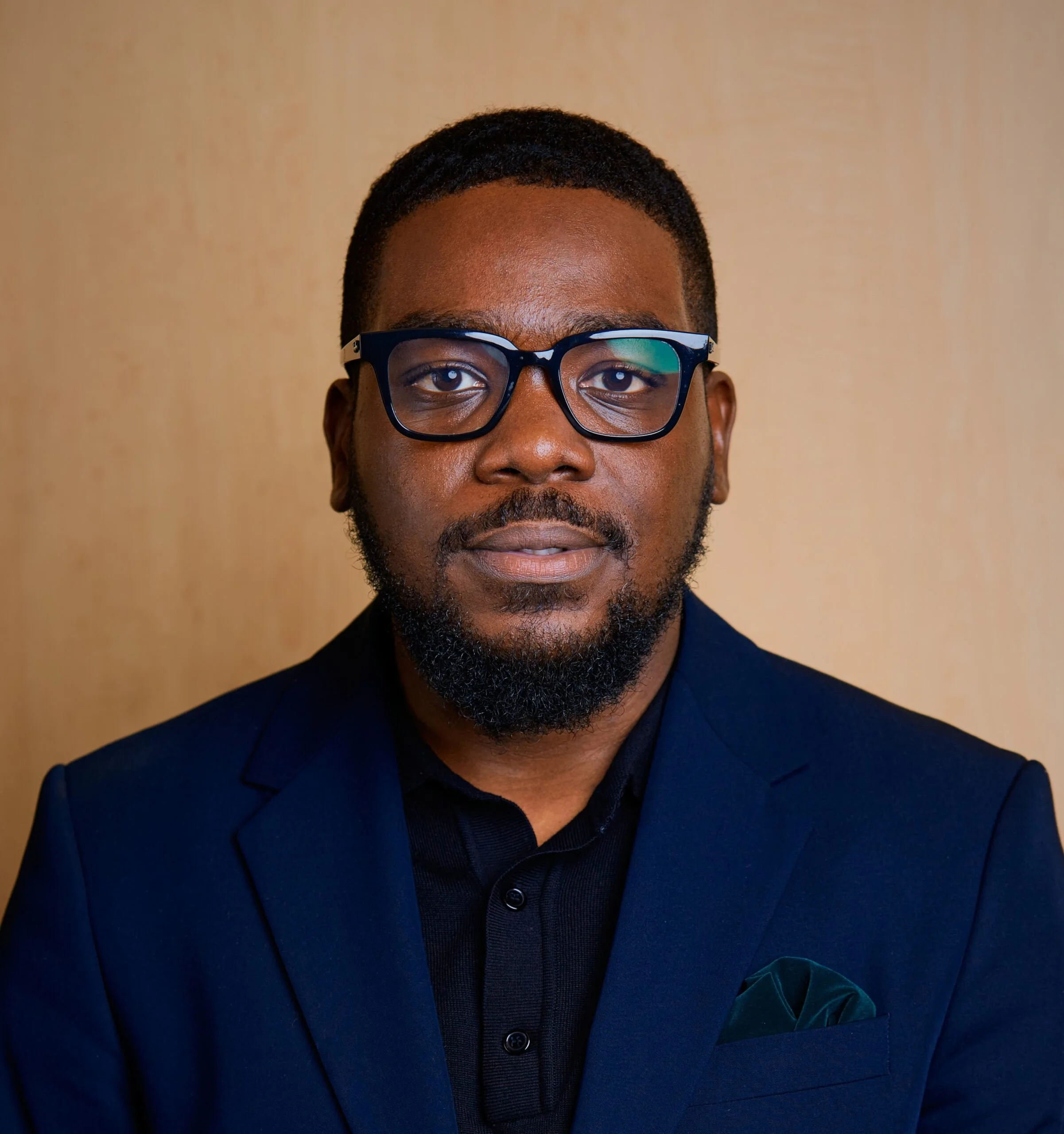 Portrait of a Black man with short hair, glasses, and a beard, wearing a dark blue suit and black shirt, standing against a wooden background.