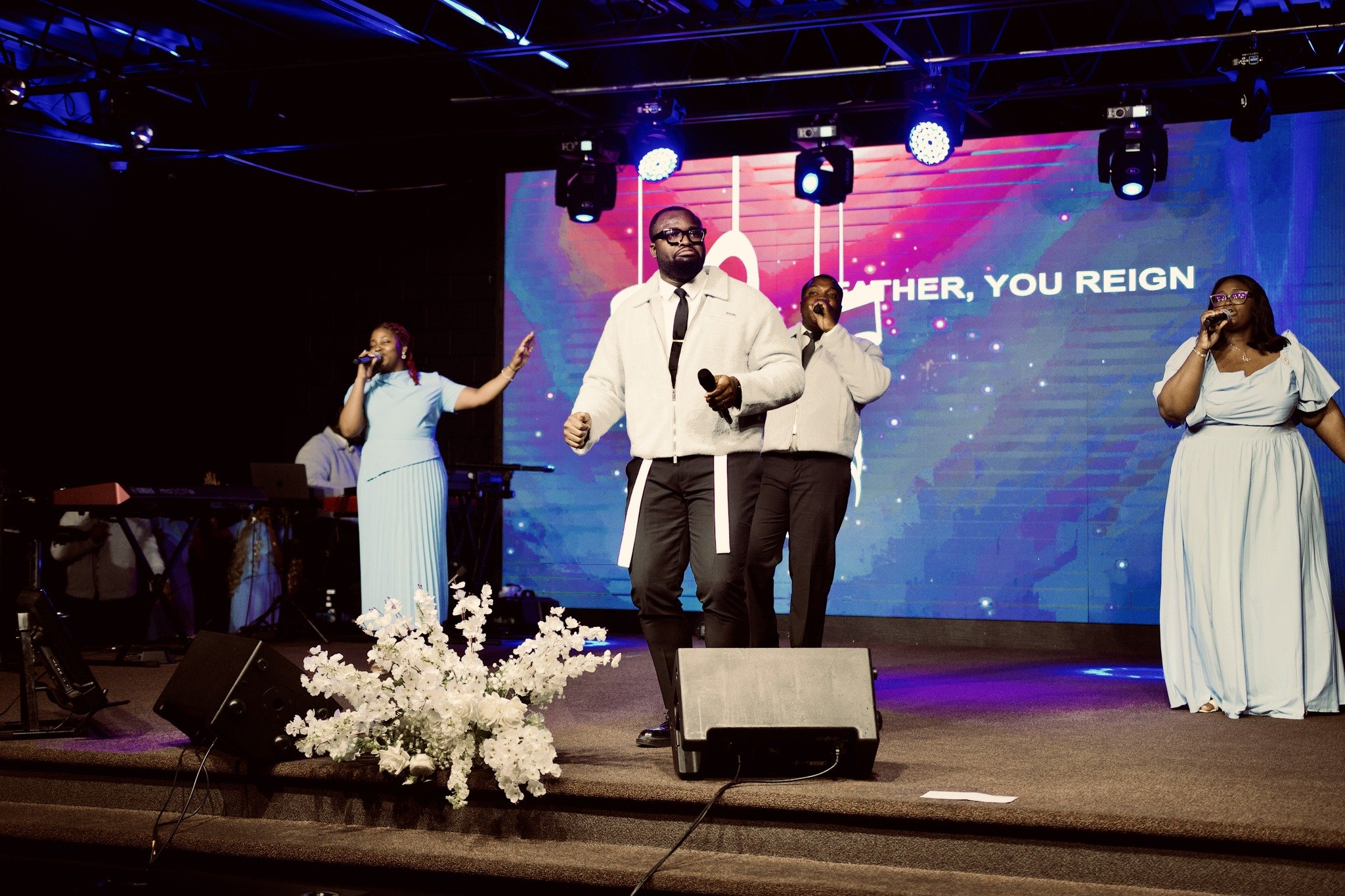 A gospel performance on stage with three singers, a keyboard player, and a large digital background screen reading 'Mother, You Reign'. The stage is decorated with white flowers and lit with blue and purple lights.