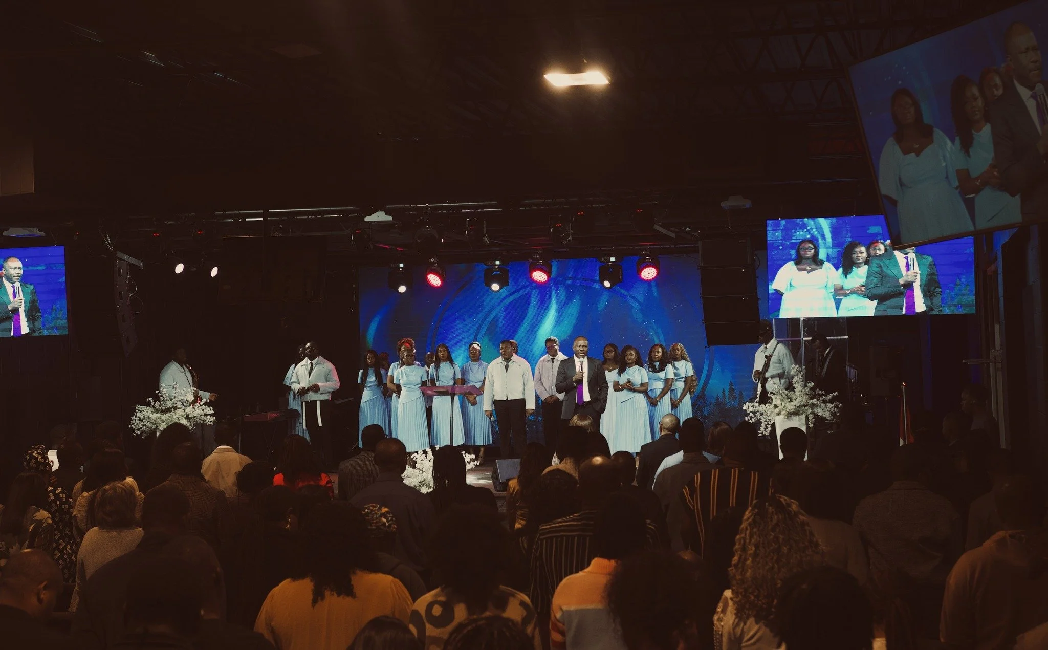 A church service or concert with a large group of people on stage, including choir members in white dresses and men in suits, with a man speaking at a microphone. The crowd is standing in front of the stage, and there are large screens showing the stage scene.