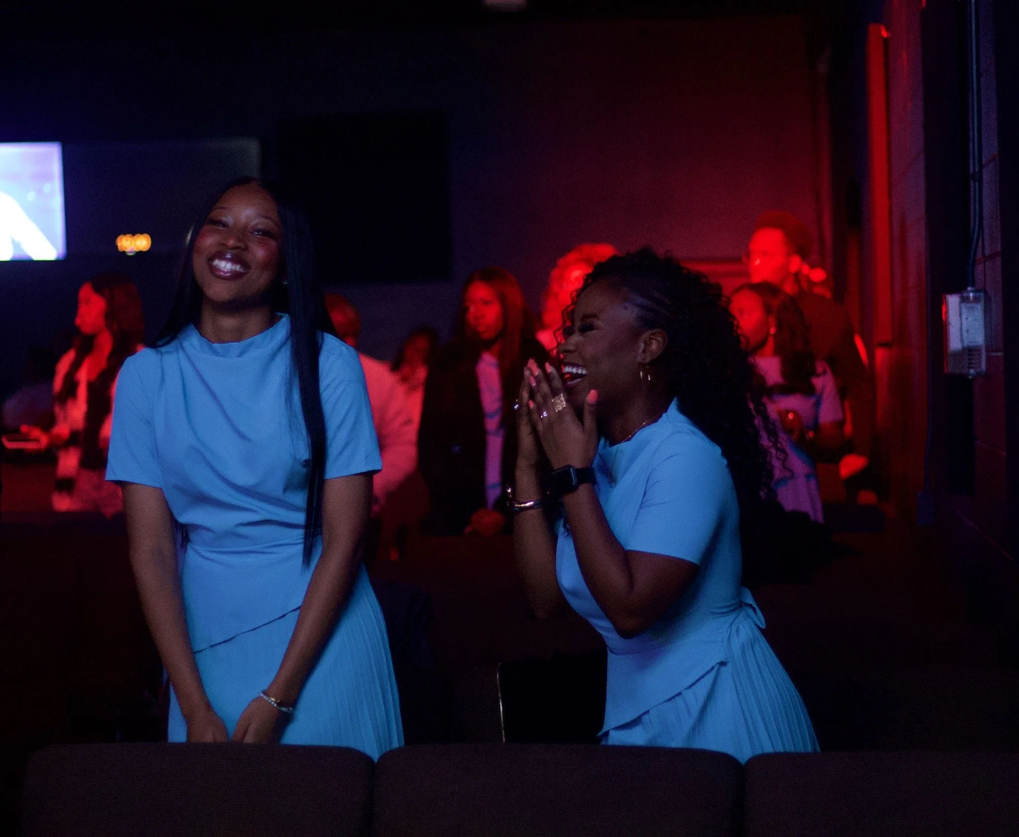 Two women in blue dresses are smiling and laughing in a dark, colorful event space with other people in the background.