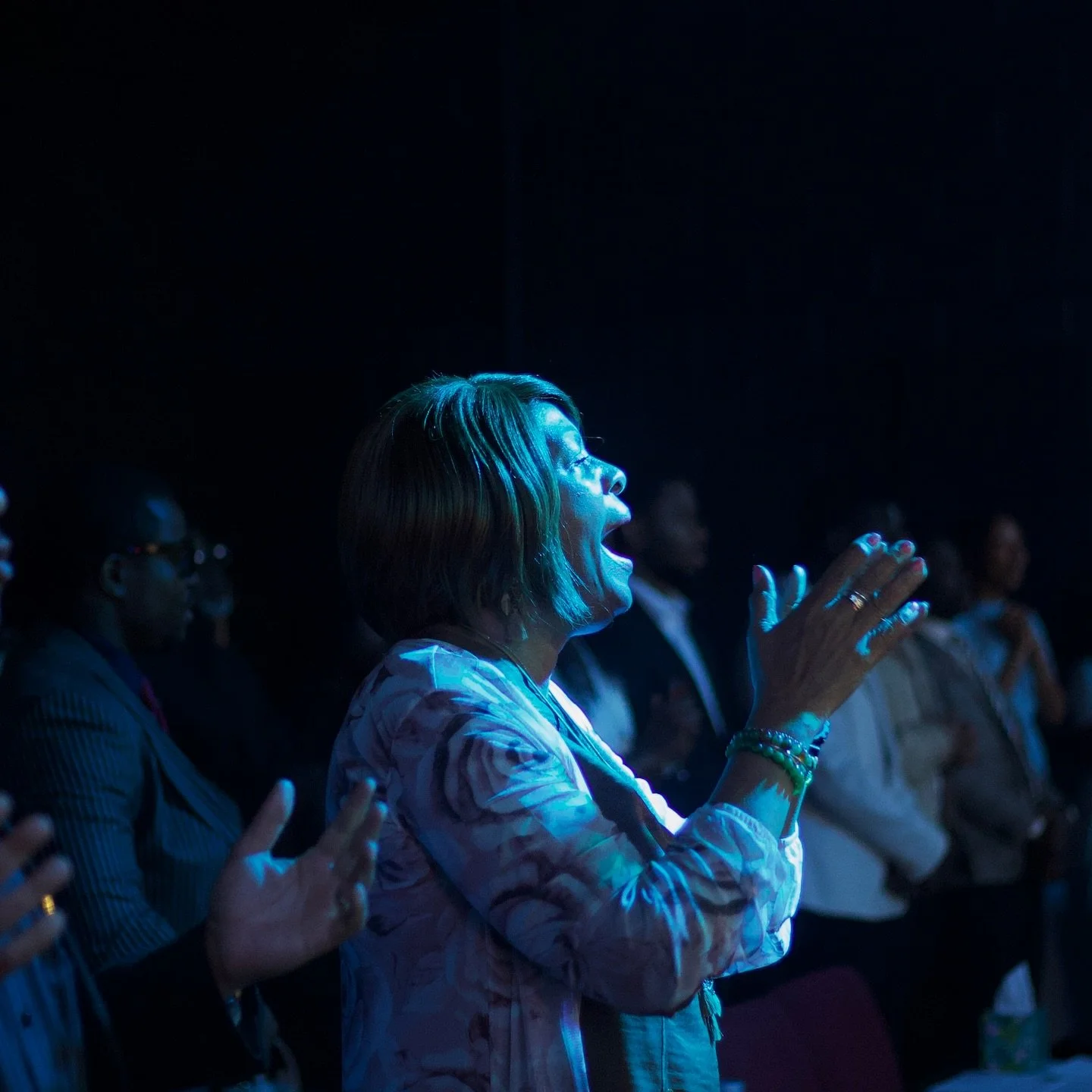 A woman with short hair claps and sings passionately at an event, illuminated by blue stage lighting, with other attendees in the background.