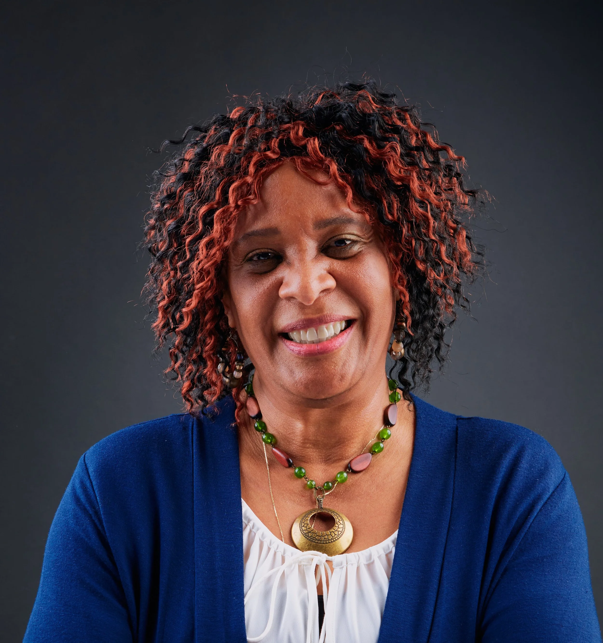 Smiling woman with curly hair, wearing a blue cardigan, white top, and green and brown necklace, against a dark background.