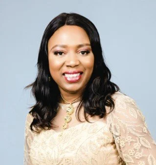 Portrait of a woman with long black hair wearing a cream lace top and gold jewelry, smiling against a light blue background.