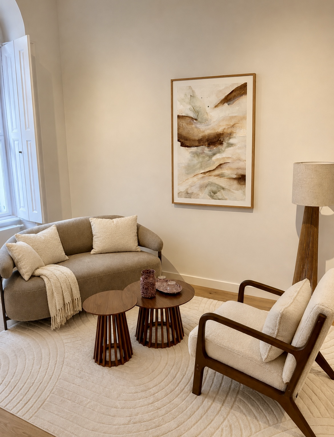 Ein Wohnzimmer mit beige Couch, beigen Sesseln, einer Stehlampe, einem abstrakten Wandbild, einem Couchtisch mit Dekor, einem Teppich und einer Fensteröffnung mit Jalousien.