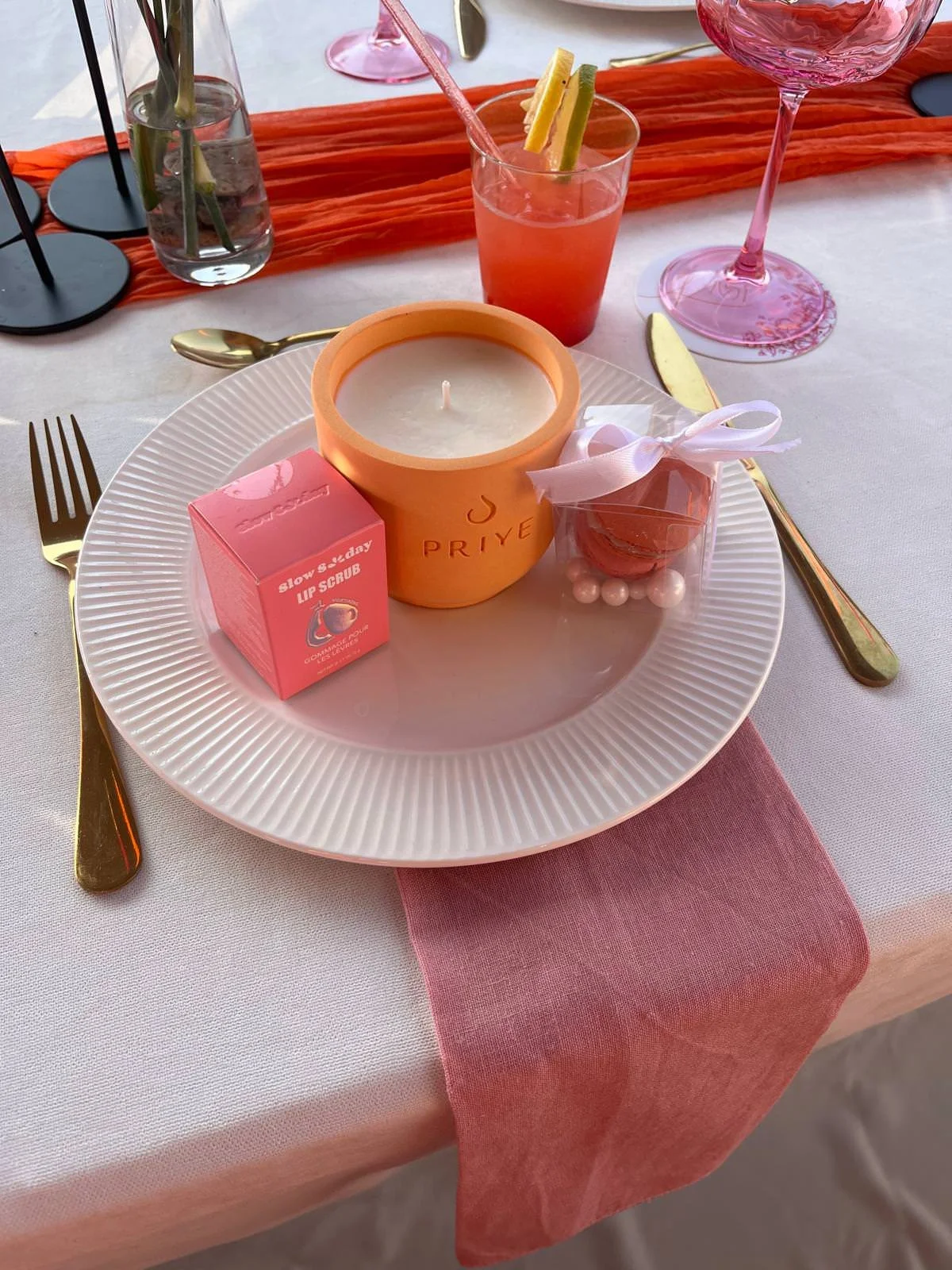 A pink-themed table setting with a white plate holding a candle, a small pink box of lip scrub, and a small pink wrapped gift with pearls. Gold utensils are placed on either side of the plate. Behind the plate are pink and clear drinks, decorative black candleholders, and a pink table runner.