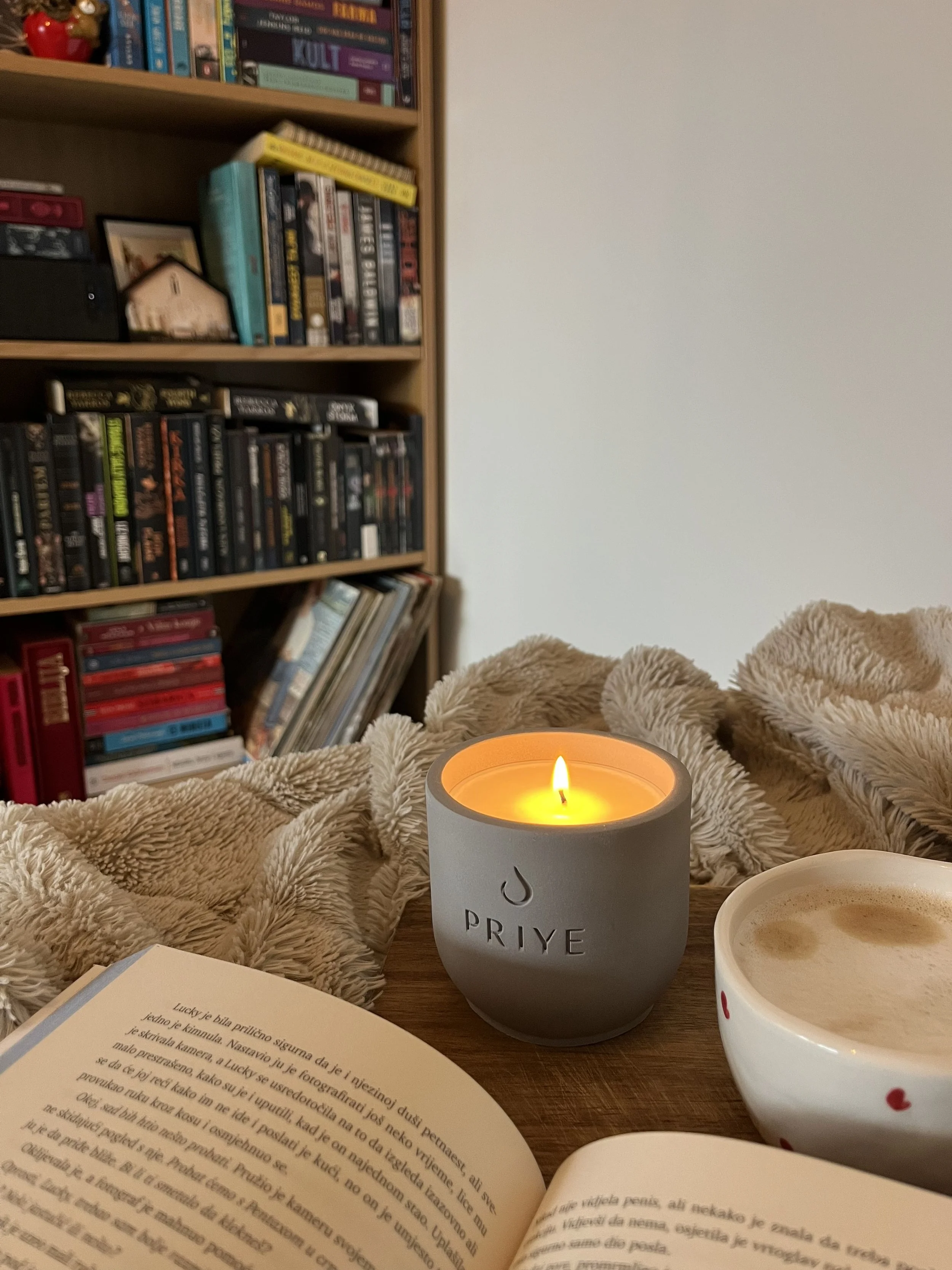 A lit candle in a gray container labeled 'PRIYE', a cup of coffee with foam, an open book, a beige blanket, and a bookshelf filled with books in the background.
