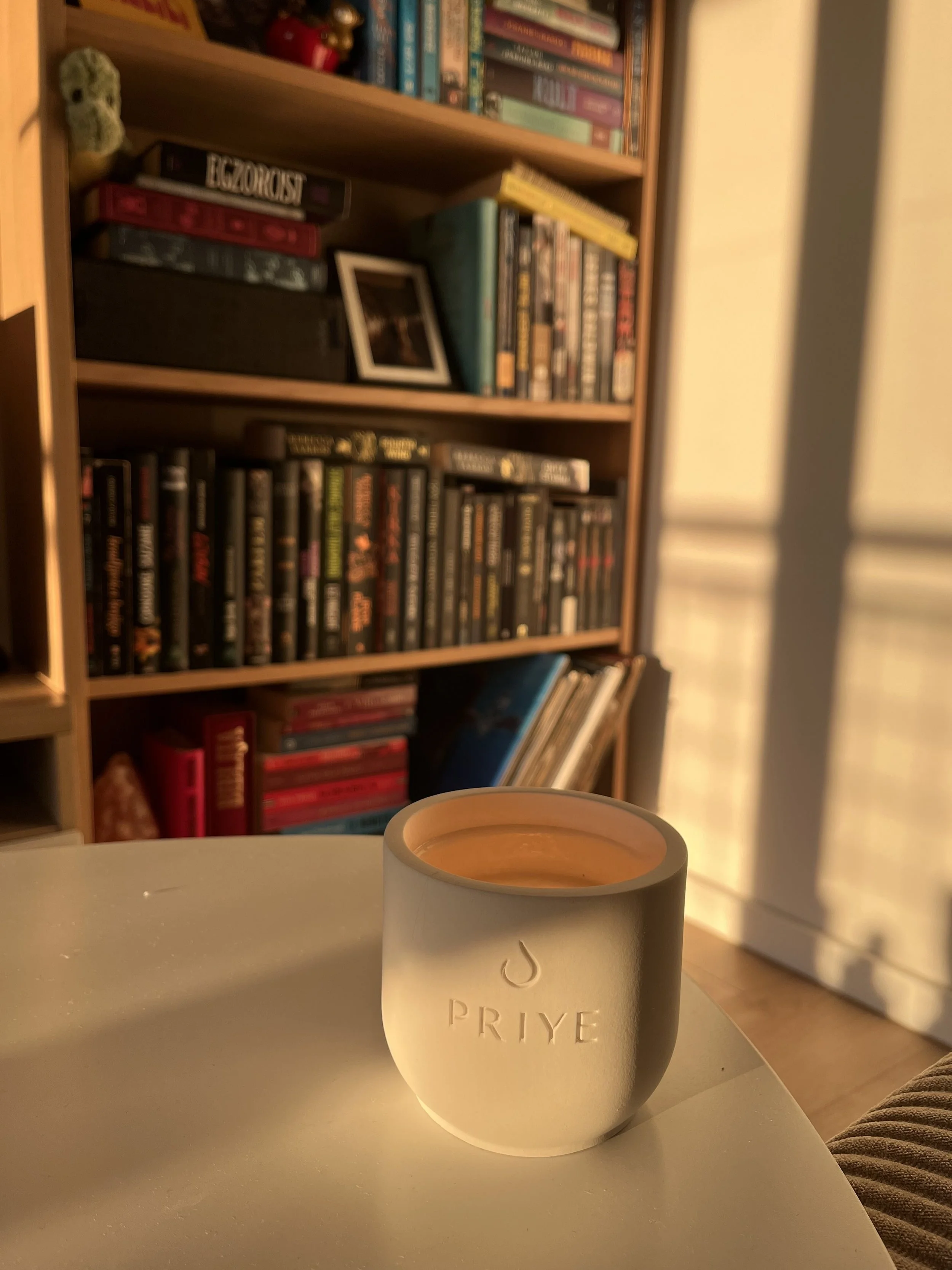 A white mug with the word 'PRIYE' and a logo is placed on a white surface, with sunlight illuminating it and a bookshelf filled with books in the background.