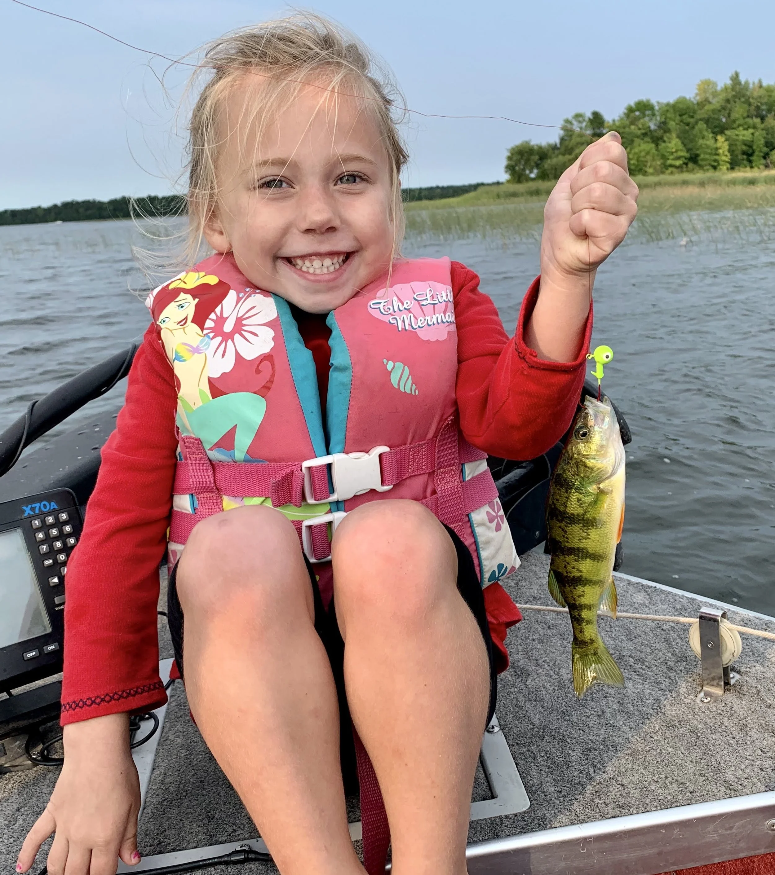 A young girl sitting in a boat on a lake, proudly holding up a perch she caught with a fishing hook. She is smiling widely, wearing a pink life jacket with Disney's The Little Mermaid design, and a red long-sleeve shirt.